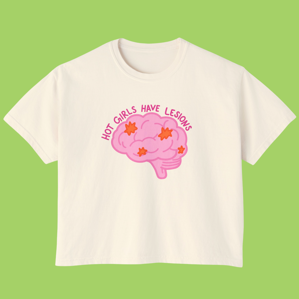 An off-white short-sleeve t-shirt with a pink cartoon brain graphic and the text 'HOT GIRLS HAVE LESIONS' in pink, displayed against a solid light green background.