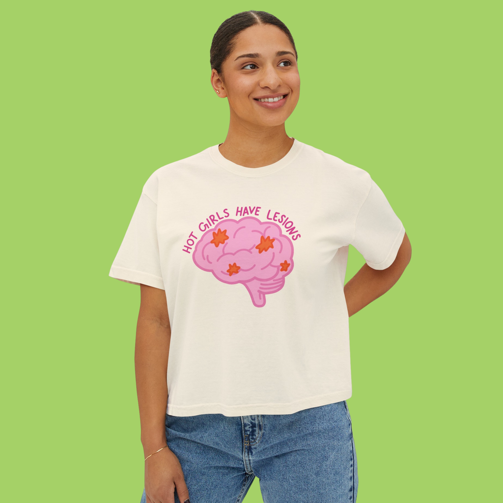 A woman wearing a white graphic t-shirt with a pink brain design and the text 'NOT GIRLS HAVE LESIONS' in pink, paired with blue jeans, set against a solid light green background.