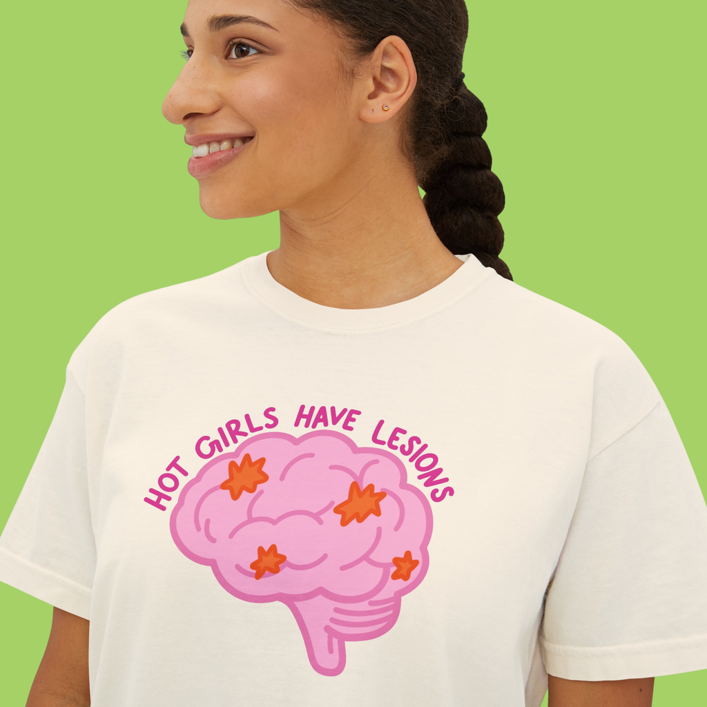 A woman wearing a white graphic t-shirt with a pink brain design and the text 'HOT GIRLS HAVE LESIONS' in pink, set against a solid light green background.