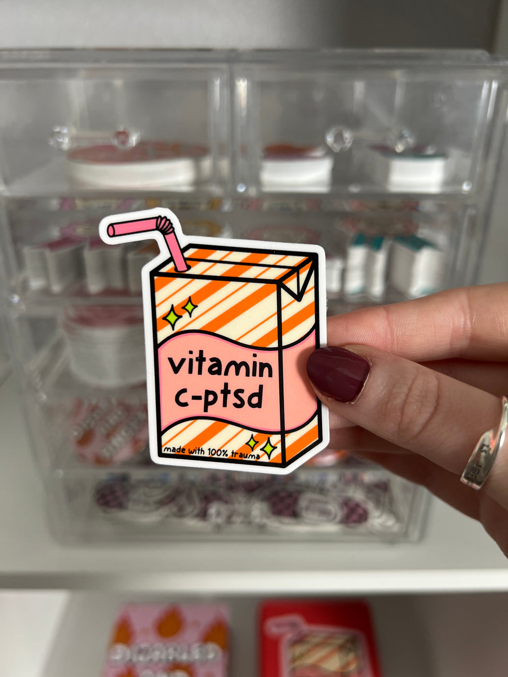 Juice box sticker, Juice box is cream with orange diagonal stripes and pink banner in the middle with yellow sparkles in either side. A pink straw is coming from the top. The black font on the juice box reads, "vitamin c-ptsd". Sticker is being held in front of product display. 