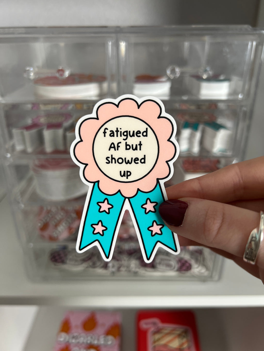 Award ribbon sticker in a light pink shade and bright turquoise with the center being cream colored. The black font reads, "fatigued AF but showed up". The sticker is being held in front of a product display.