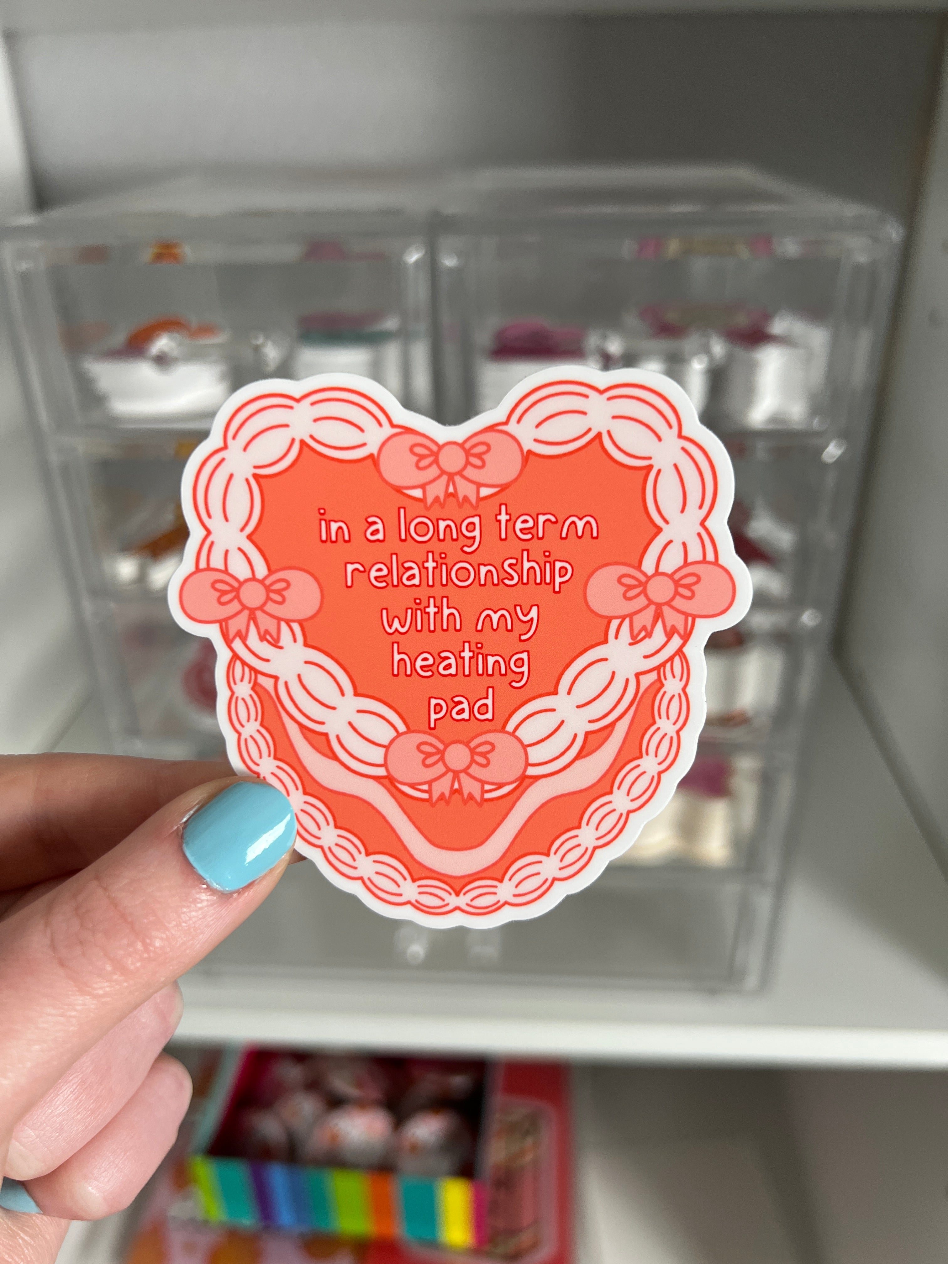 A coral heart-shaped vinyl sticker with the words "in a long term relationship with my heating pad" in white font outlined in red The sticker has a decorative border with bows and a scalloped edge. The sticker is being held against a clear storage container.