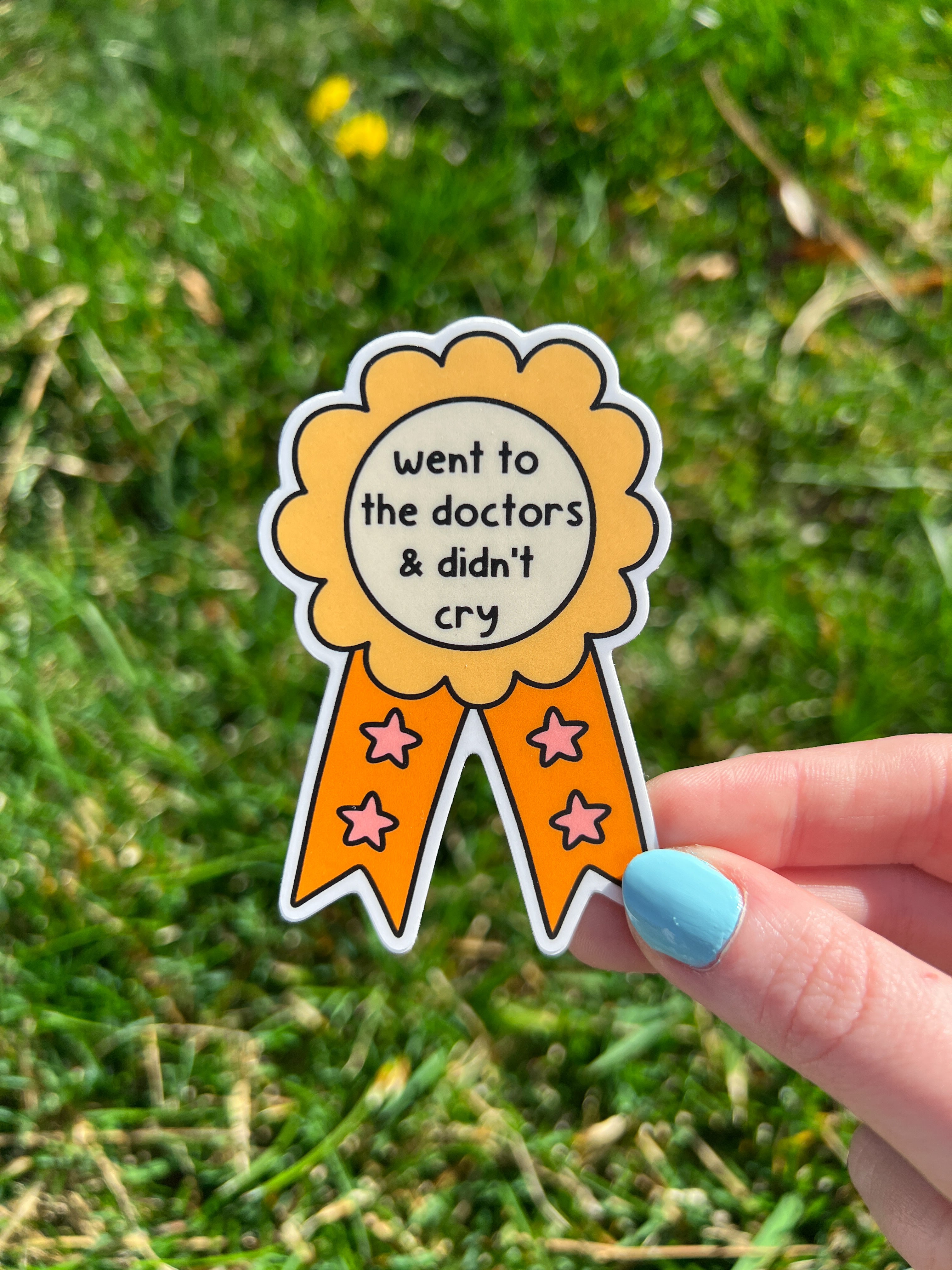 A sticker in the shape of a rosette award ribbon with the text "went to the doctors & didn't cry". The rosette is yellow with orange accents and orange ribbons with pink stars.