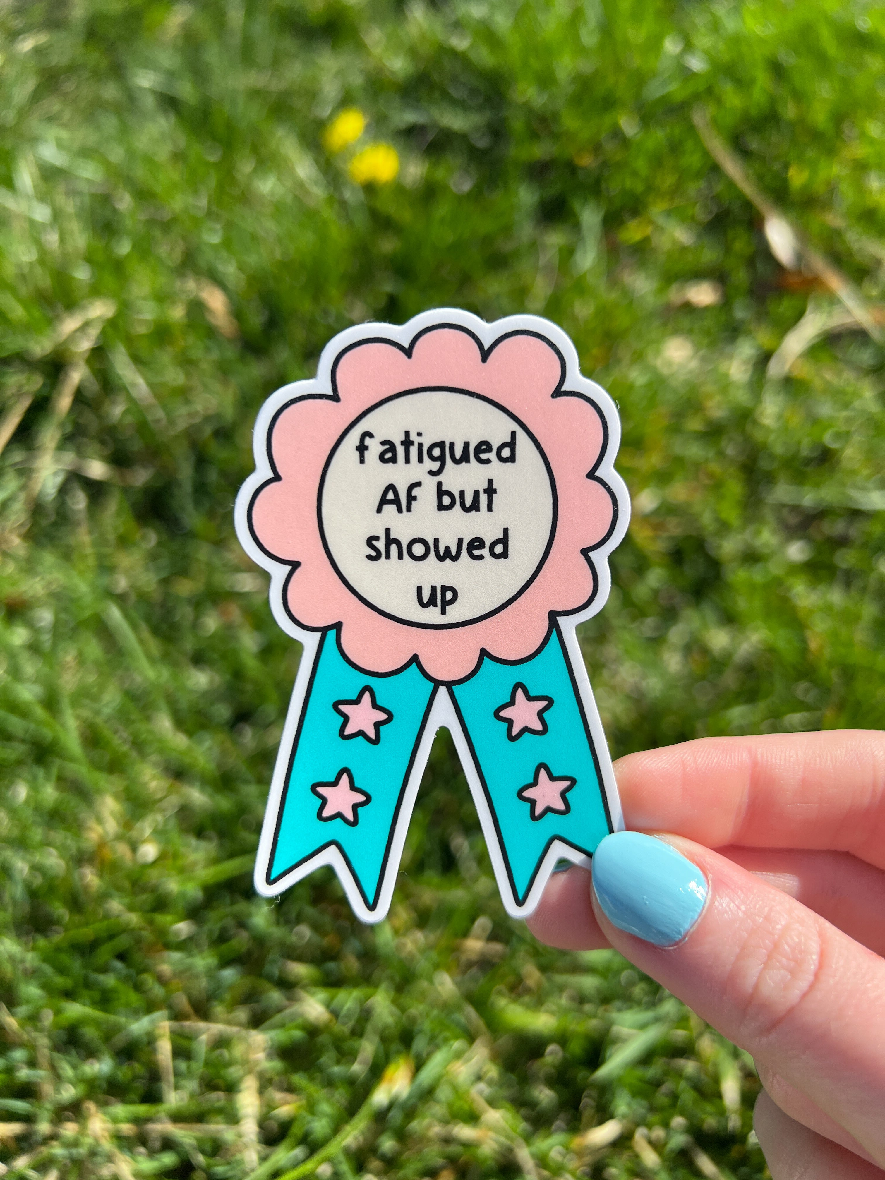 Award ribbon sticker in a light pink shade and bright turquoise with the center being cream colored. The black font reads, "fatigued AF but showed up". The sticker is being held over grass.
