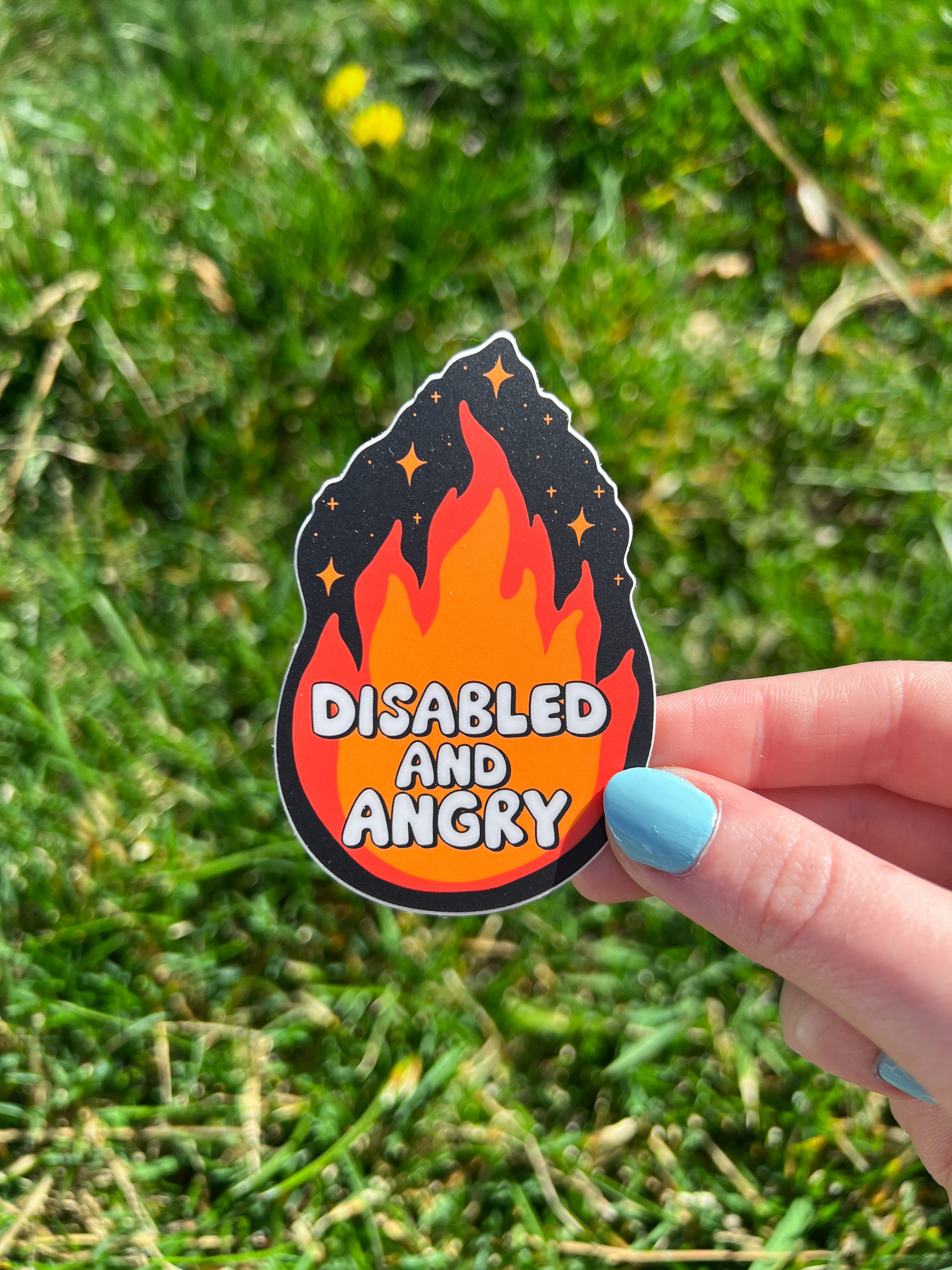 A die-cut sticker featuring a stylized flame graphic in shades of orange and red against a dark background with small star accents. The text "DISABLED AND ANGRY" is printed in a bold, white, rounded font across the center of the flame.