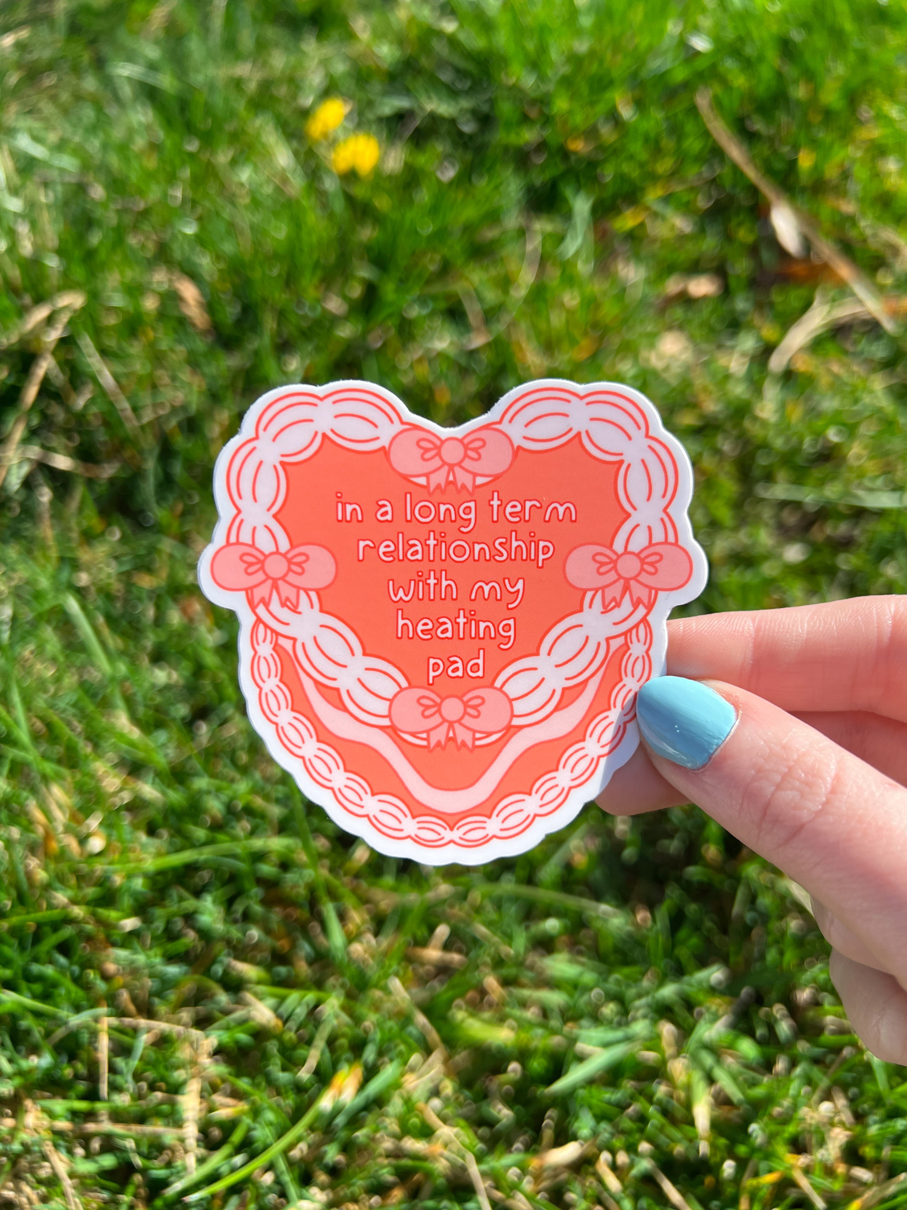 A coral heart-shaped vinyl sticker with the words "in a long term relationship with my heating pad" in white font outlined in red The sticker has a decorative border with bows and a scalloped edge. The sticker is being held against a grassy background. 
