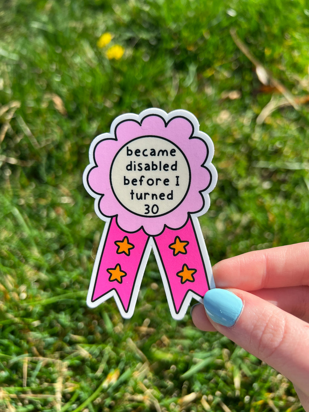 A die-cut sticker shaped like a rosette award ribbon. The center is a cream circle with the black text 'became disabled before I turned 30'. It has a pink scalloped border and two tails in hot pink with orange stars.