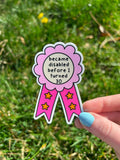 A die-cut sticker shaped like a rosette award ribbon. The center is a cream circle with the black text 'became disabled before I turned 30'. It has a pink scalloped border and two tails in hot pink with orange stars.