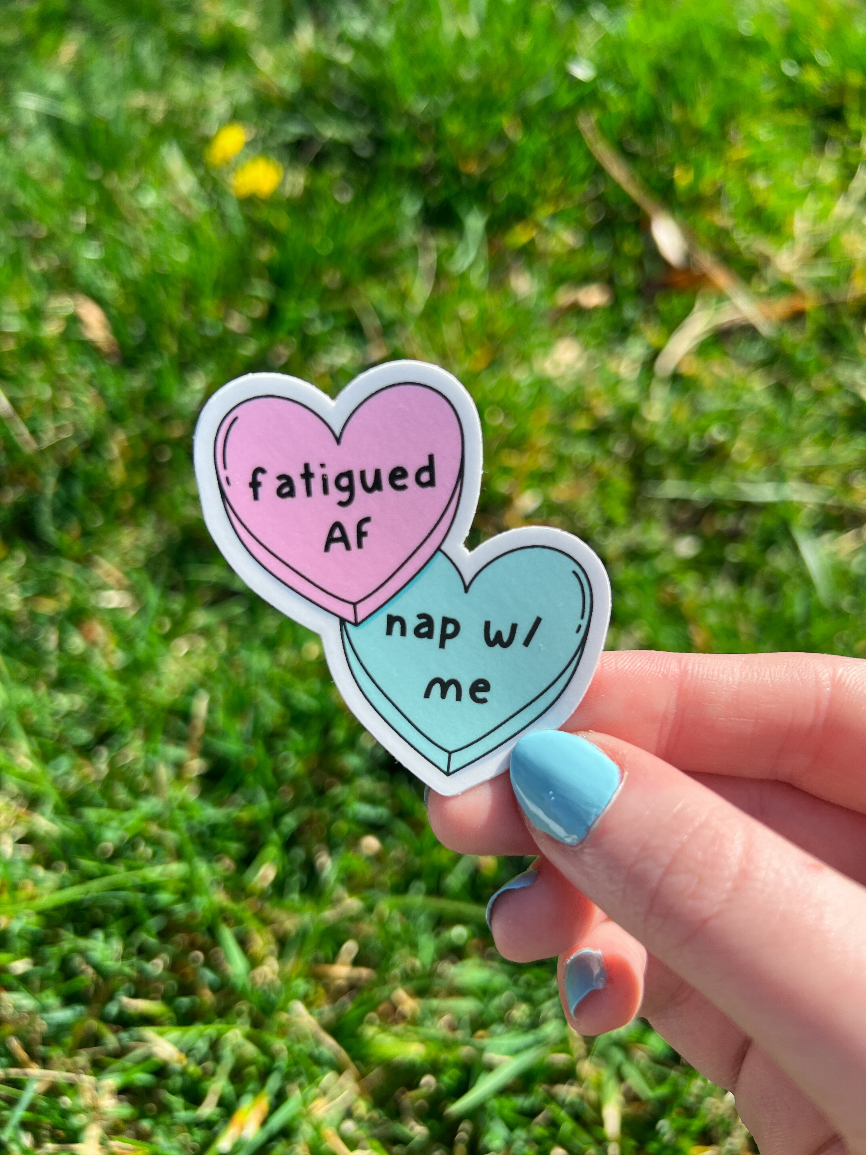 Candy heart sticker, one heart is in light pink and the other is in light blue. The font on the pink one reads, "fatigued AF" while the blue heart reads, "nap with me." Sticker is being held over grass on a sunny day. 