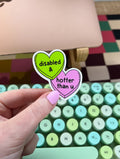 Two heart-shaped vinyl sticker. One is lime green with the words "disabled &" in a black font. The other is pink with the words "hotter than u" in a black font. The sticker is held over a keyboard.