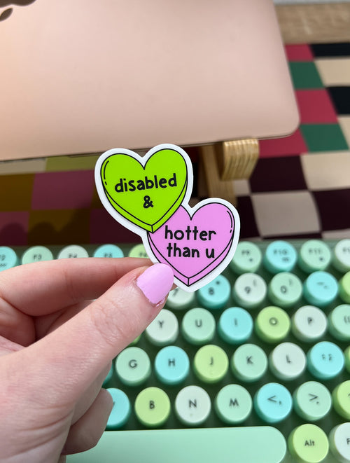 Two heart-shaped vinyl sticker. One is lime green with the words "disabled &" in a black font. The other is pink with the words "hotter than u" in a black font. The sticker is held over a keyboard.