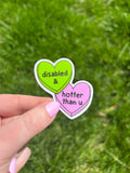 Two heart-shaped vinyl sticker. One is lime green with the words "disabled &" in a black font. The other is pink with the words "hotter than u" in a black font. The stickers are held against a green grass background.
