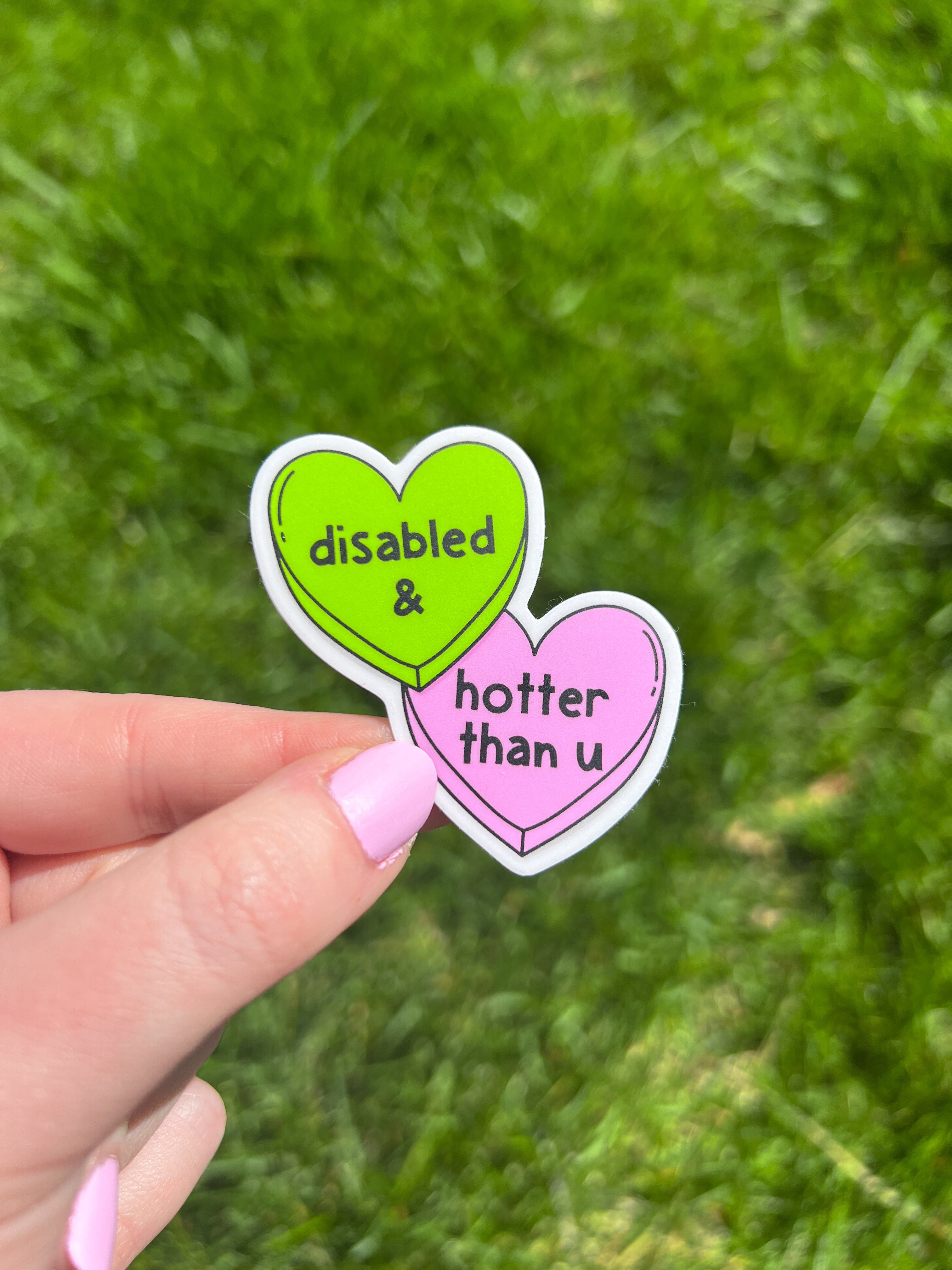 Two heart-shaped vinyl sticker. One is lime green with the words "disabled &" in a black font. The other is pink with the words "hotter than u" in a black font. The stickers are held against a green grass background.
