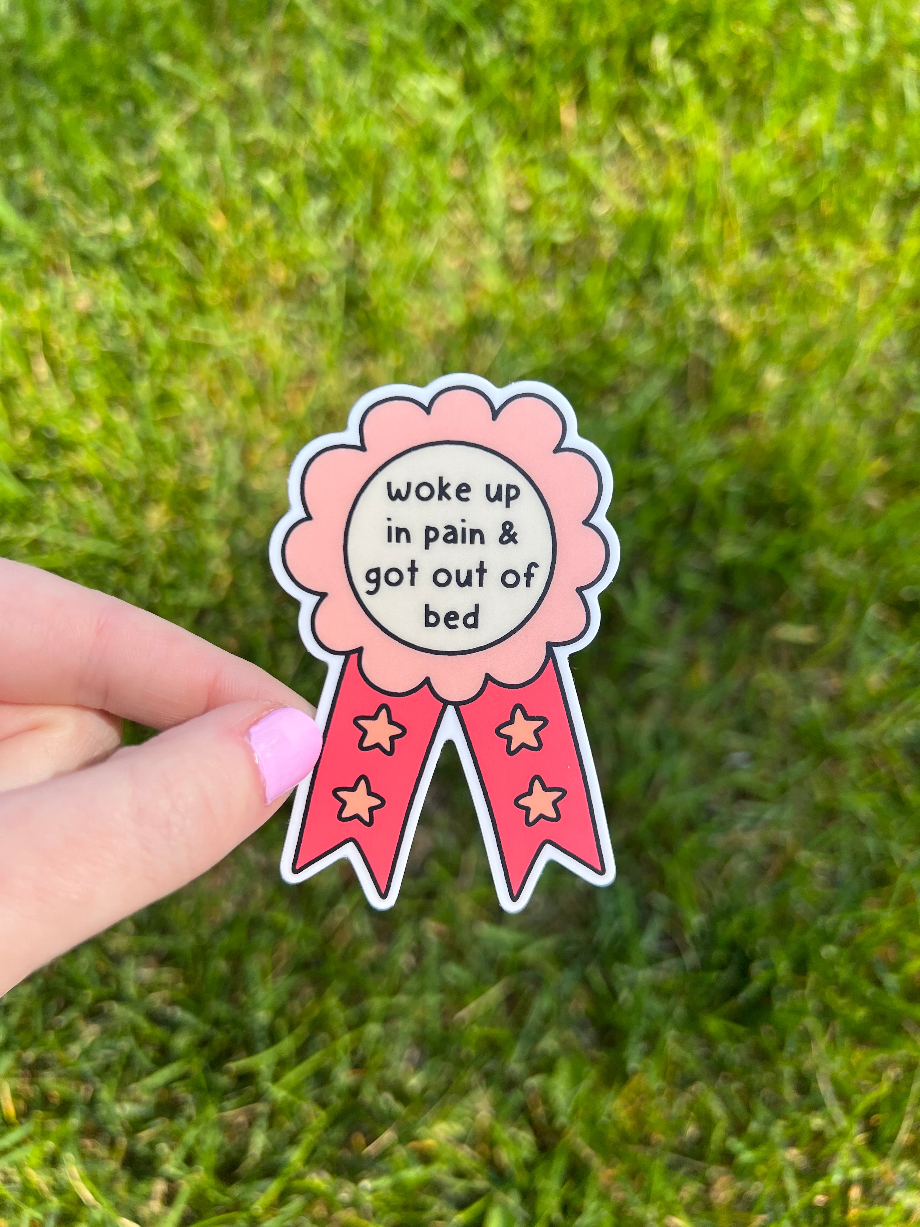 A sticker with a pink and peach rosette design and red ribbons. The text "woke up in pain & got out of bed" is in the center. Four gold stars are on the ribbons. The sticker is held against a green grassy background.