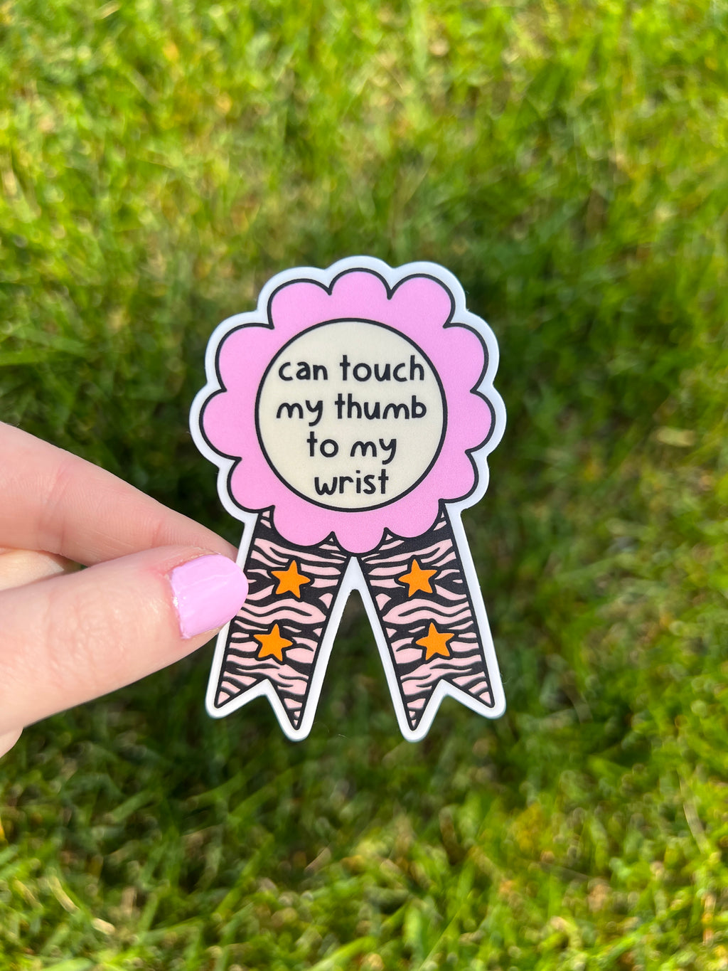 A vinyl die-cut sticker shaped like a rosette award ribbon. The center is a cream circle with the black text 'can touch my thumb to my wrist'. It has a pink scalloped border and two tails featuring a black zebra print with orange stars.