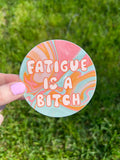 A circular sticker featuring a swirling marbled pattern in shades of pink, orange, and light blue. The text "FATIGUE IS A BITCH" is printed in the center in a bold, rounded, light-colored font with a thin orange outline.