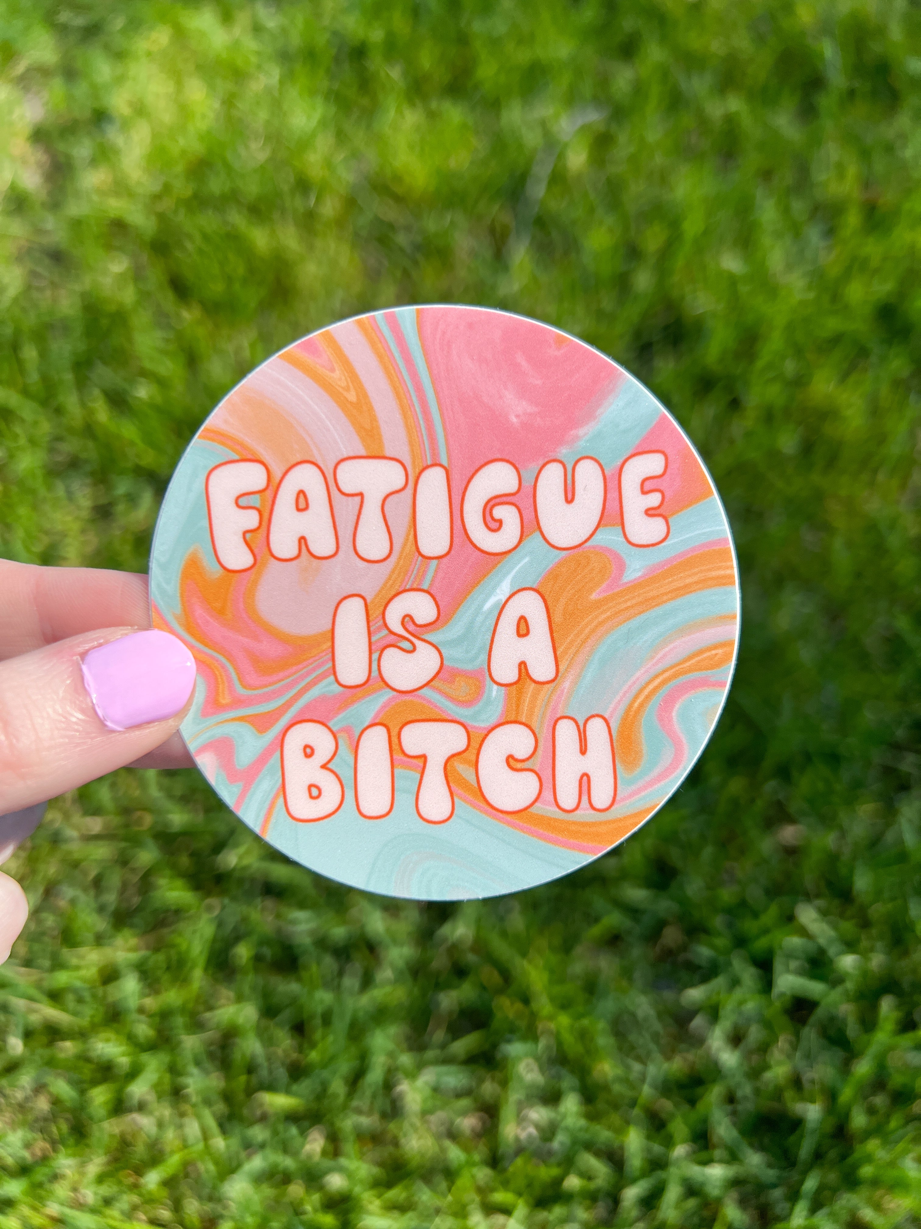 A circular sticker featuring a swirling marbled pattern in shades of pink, orange, and light blue. The text "FATIGUE IS A BITCH" is printed in the center in a bold, rounded, light-colored font with a thin orange outline.