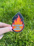 A die-cut sticker featuring a stylized flame graphic in shades of orange and red against a dark background with small star accents. The text "DISABLED AND ANGRY" is printed in a bold, white, rounded font across the center of the flame.