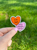 Two heart-shaped stickers with text. The top sticker is orange with the words "I'm hot" in black. The bottom sticker is pink with the words "& my stomach hurts" in black. The stickers are held by a hand with blue nail polish.