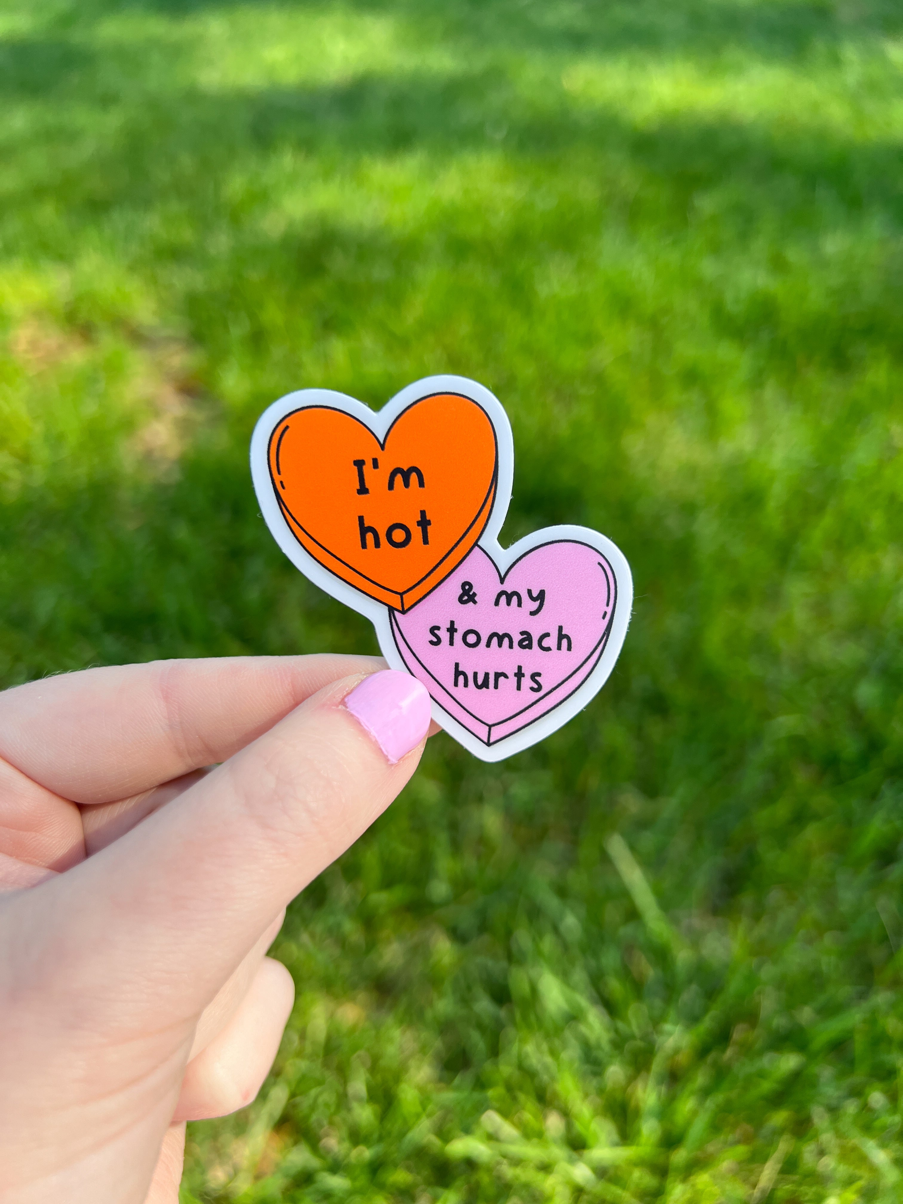 Two heart-shaped stickers with text. The top sticker is orange with the words "I'm hot" in black. The bottom sticker is pink with the words "& my stomach hurts" in black. The stickers are held by a hand with blue nail polish.