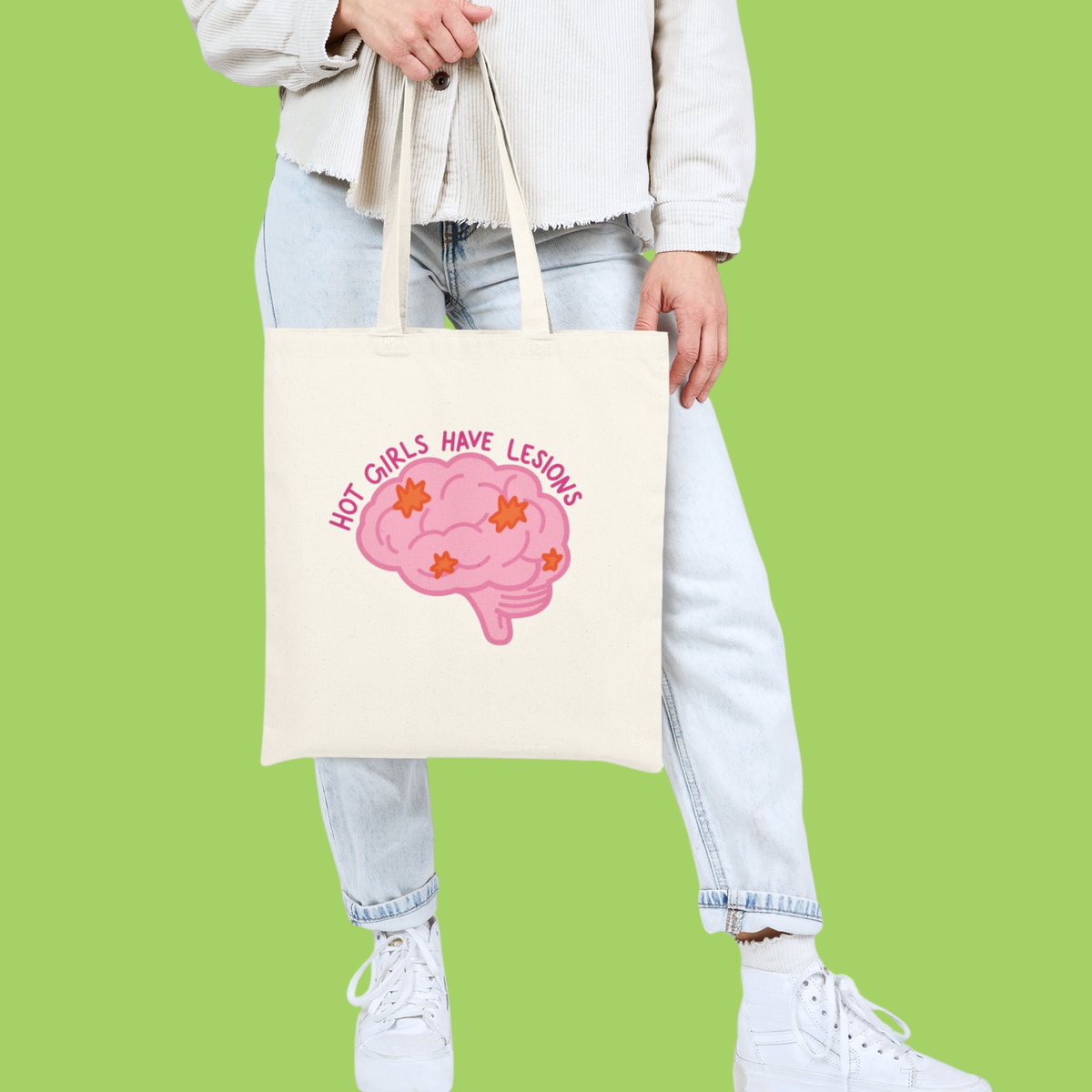 A person wearing a white jacket, light blue jeans, and white sneakers holds a cream canvas tote bag with a pink brain graphic and the text 'HOT GIRLS HAVE LESIONS' against a solid light green background.