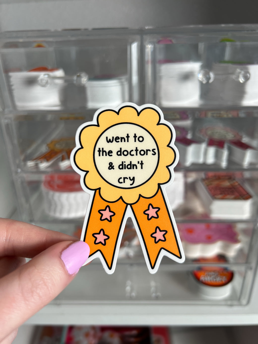 A sticker in the shape of a rosette award ribbon with the text "went to the doctors & didn't cry". The rosette is yellow with orange accents and orange ribbons with pink stars.