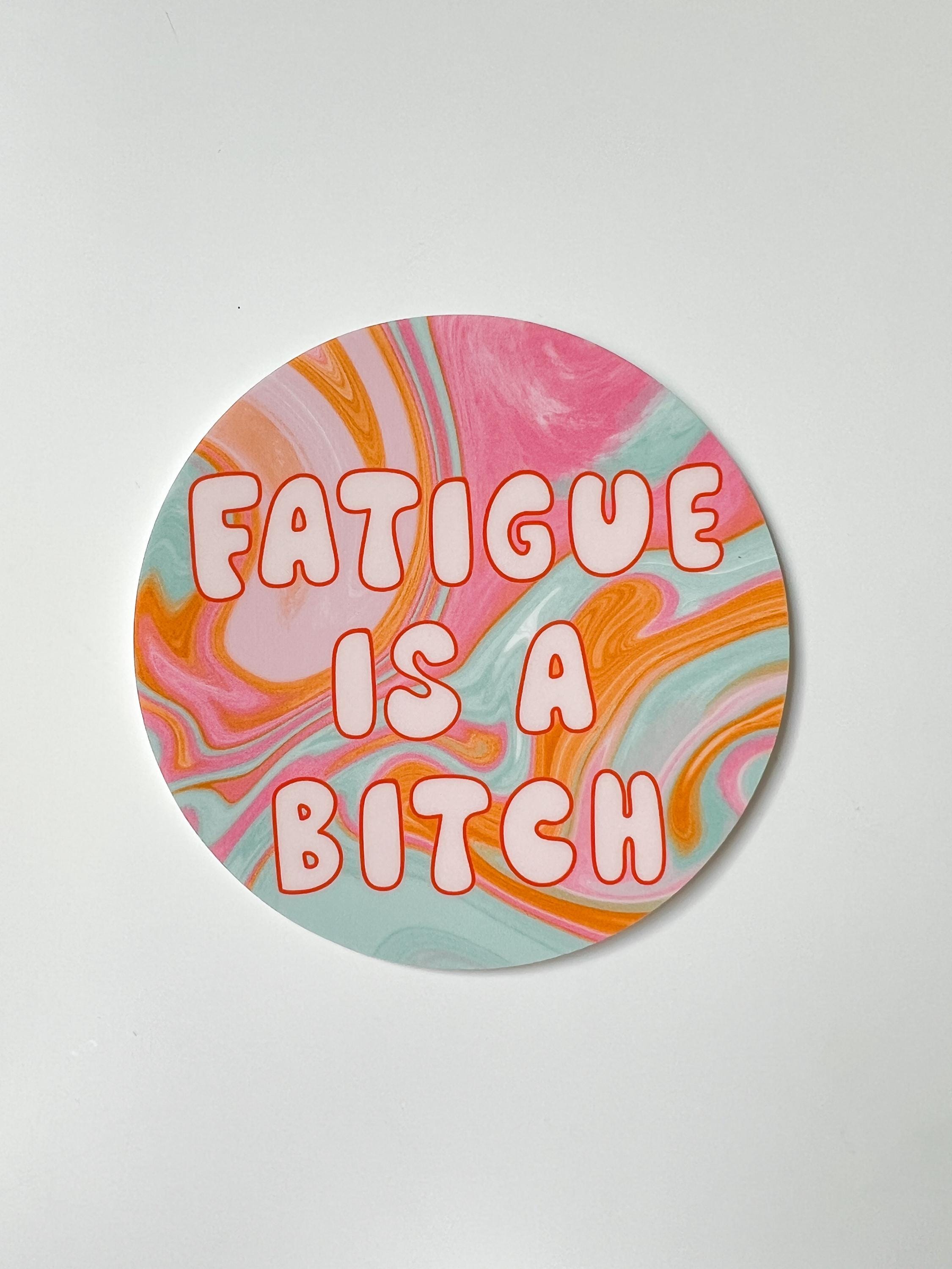 Round sticker in tie dye colors of pink, yellow orange, light blue, and white with large bubble letters outlined in burnt orange and filled in with a cream color reading, "fatigue is a bitch". Sticker is laid on a white table.