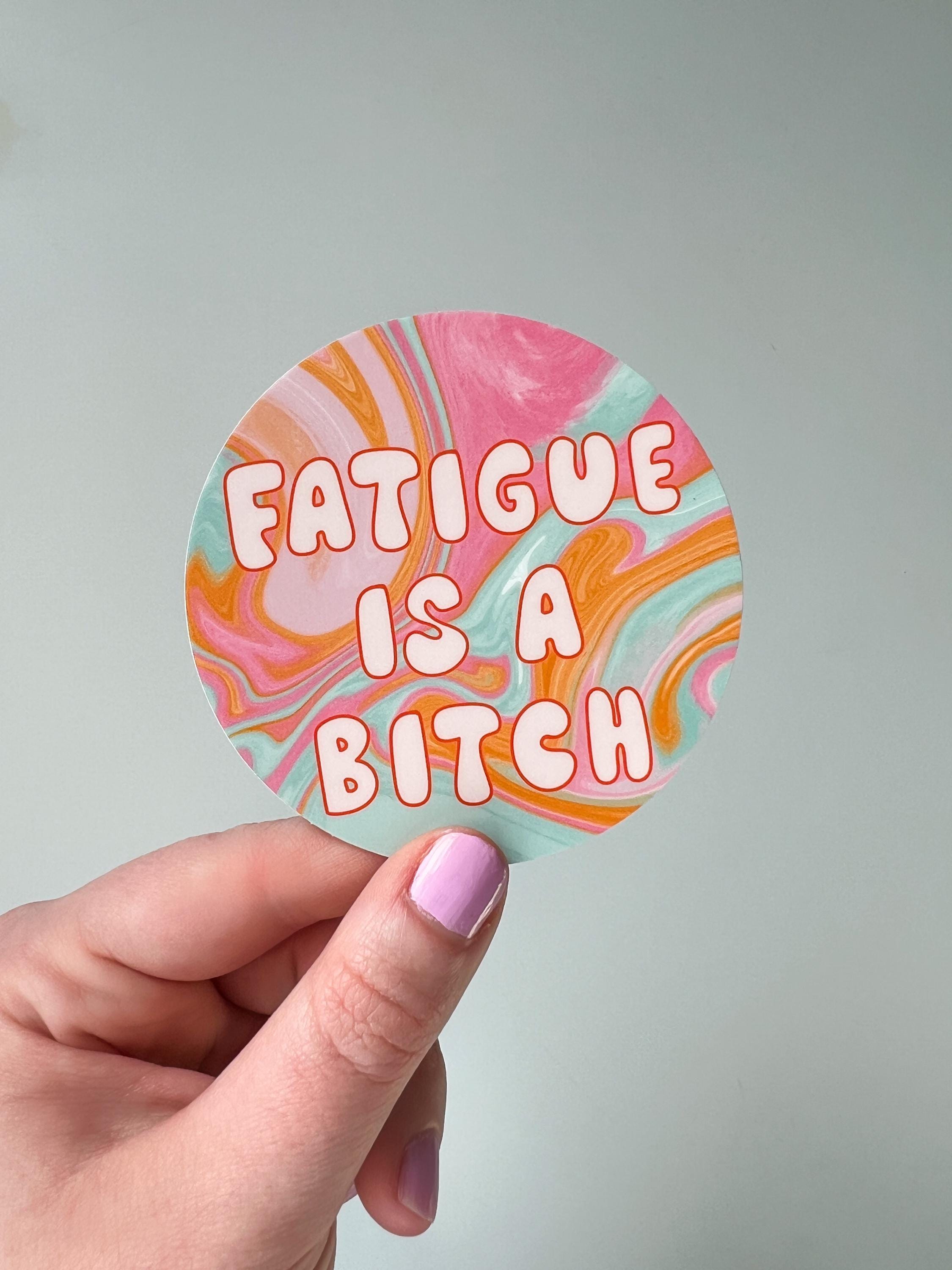 Round sticker in tie dye colors of pink, yellow orange, light blue, and white with large bubble letters outlined in burnt orange and filled in with a cream color reading, "fatigue is a bitch". Sticker is being held in front of white wall. 
