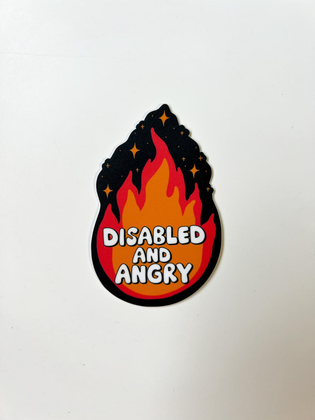 Flame shaped sticker with black background, an orange and red flame with sparkles around the top. The font in the flames center reads, "disabled and angry". The sticker is laid on white table.