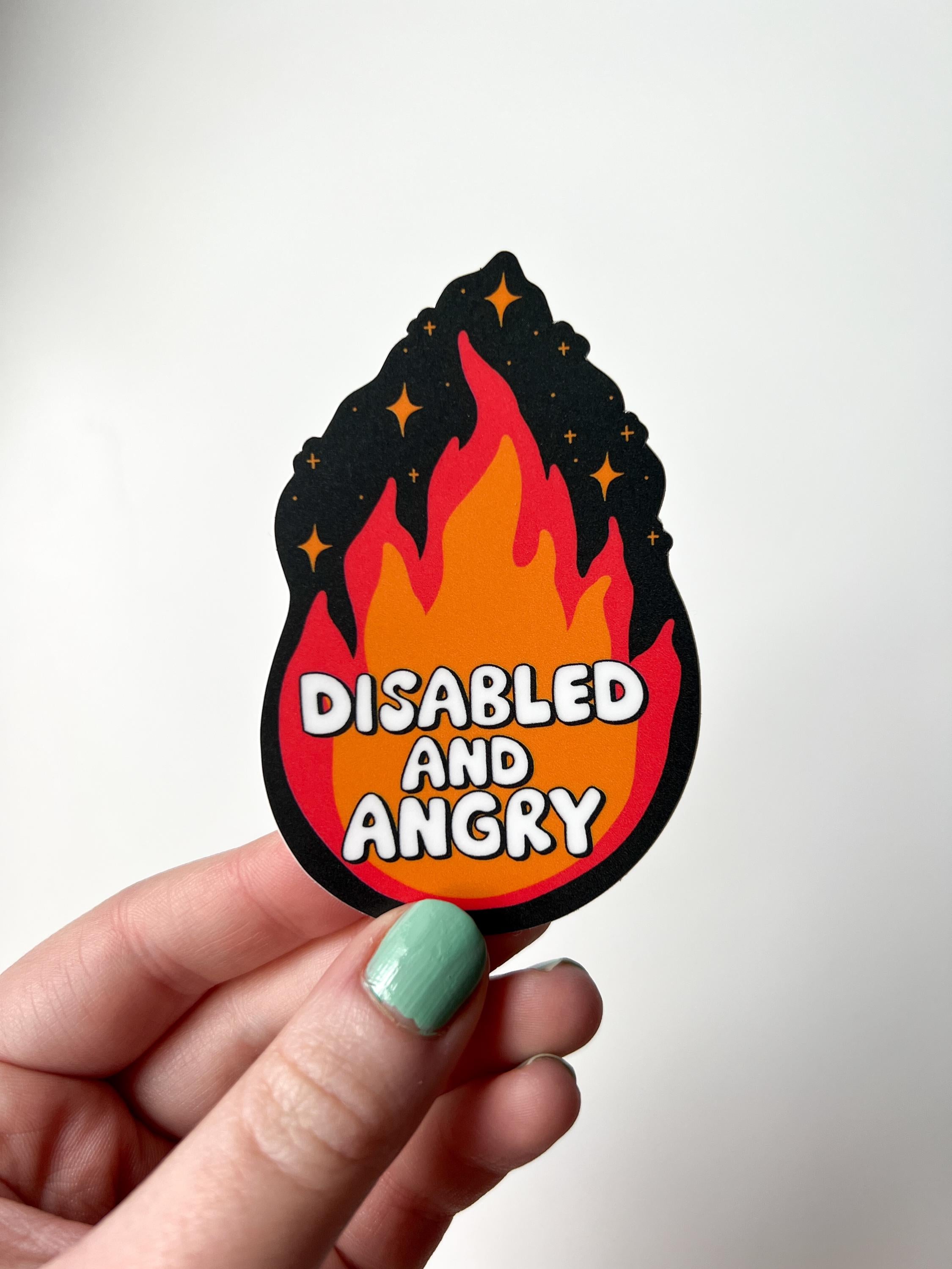 Flame shaped sticker with black background, an orange and red flame with sparkles around the top. The font in the flames center reads, "disabled and angry". The sticker is held over white table. 