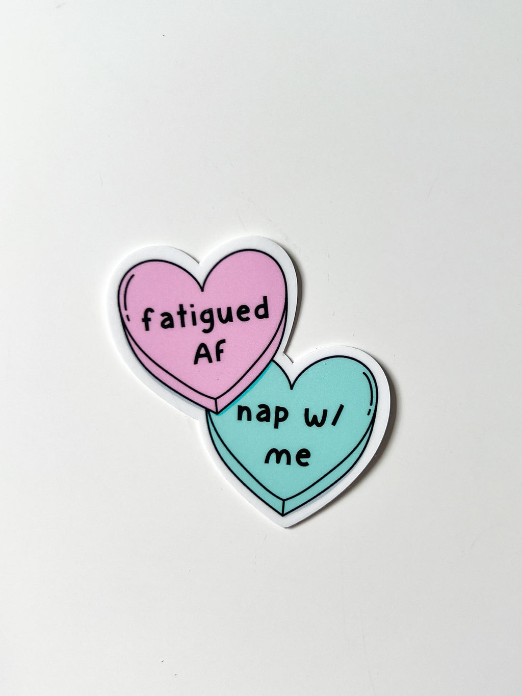 Candy heart sticker, one heart is in light pink and the other is in light blue. The font on the pink one reads, "fatigued AF" while the blue heart reads, "nap with me." Sticker is laid on white table. 