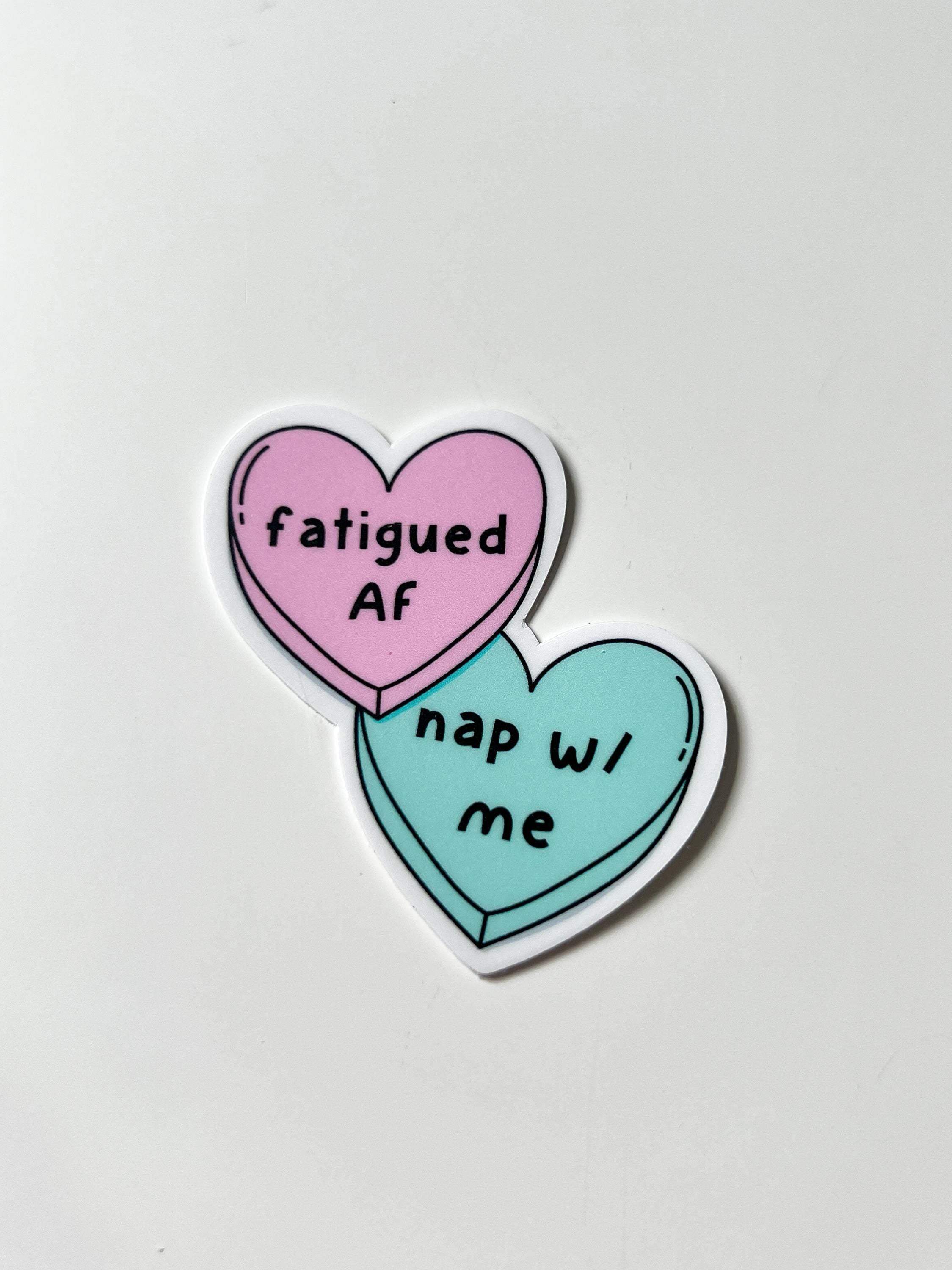 Candy heart sticker, one heart is in light pink and the other is in light blue. The font on the pink one reads, "fatigued AF" while the blue heart reads, "nap with me." Sticker is laid on white table. 
