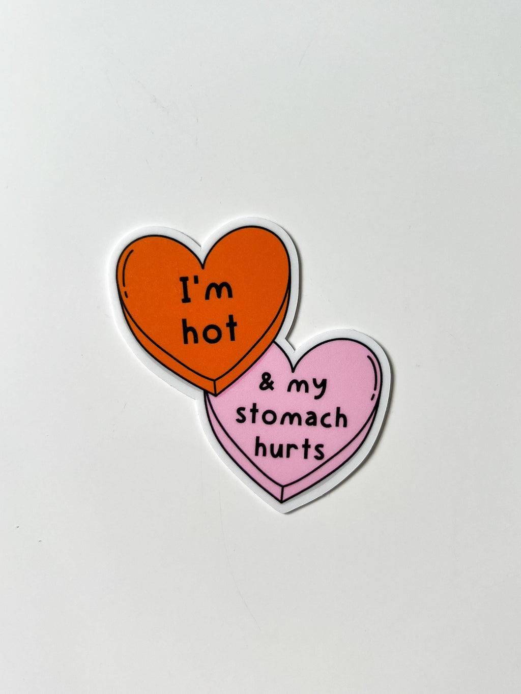 Candy heart sticker with one heart being a bright shade of orange and the other in light pink. The bright orange heart reads, "I'm hot" while the light prink heart reads, "and my stomach hurts". Sticker is laid on white table.