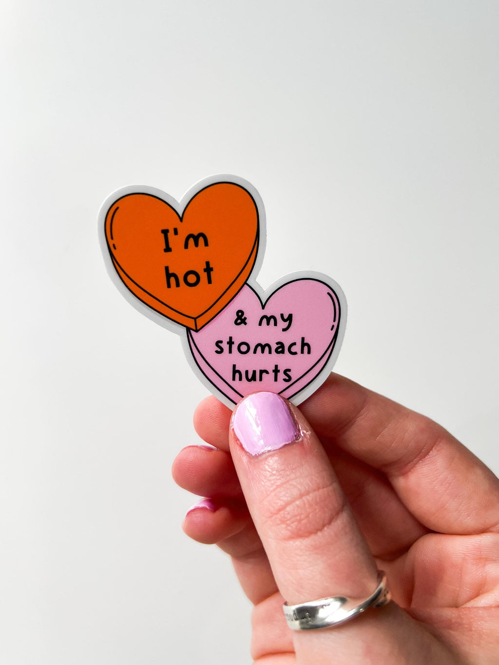 Candy heart sticker with one heart being a bright shade of orange and the other in light pink. The bright orange heart reads, "I'm hot" while the light prink heart reads, "and my stomach hurts". Sticker is held in front of white table.