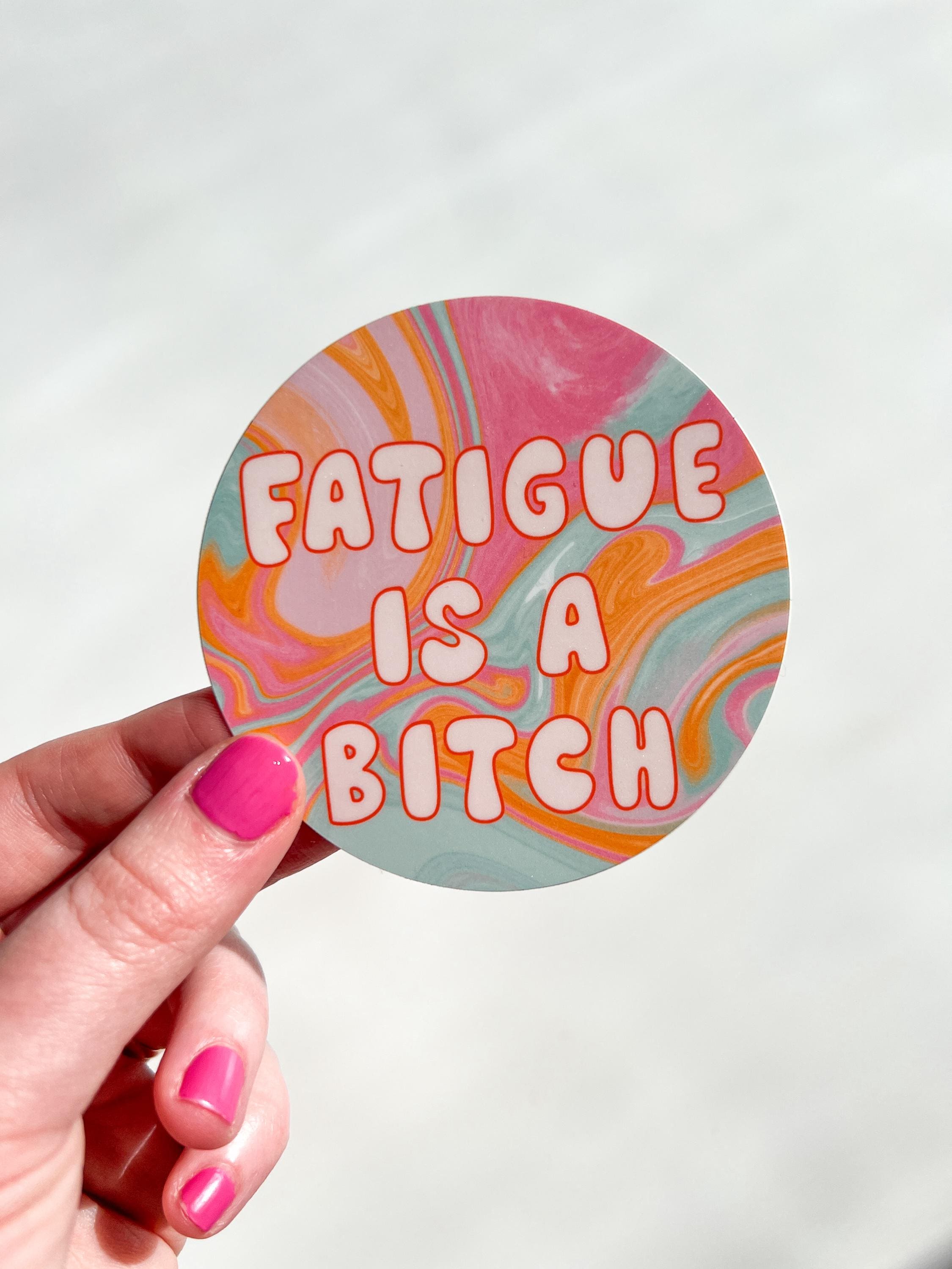 Round sticker in tie dye colors of pink, yellow orange, light blue, and white with large bubble letters outlined in burnt orange and filled in with a cream color reading, "fatigue is a bitch". Sticker is held in front of white table.