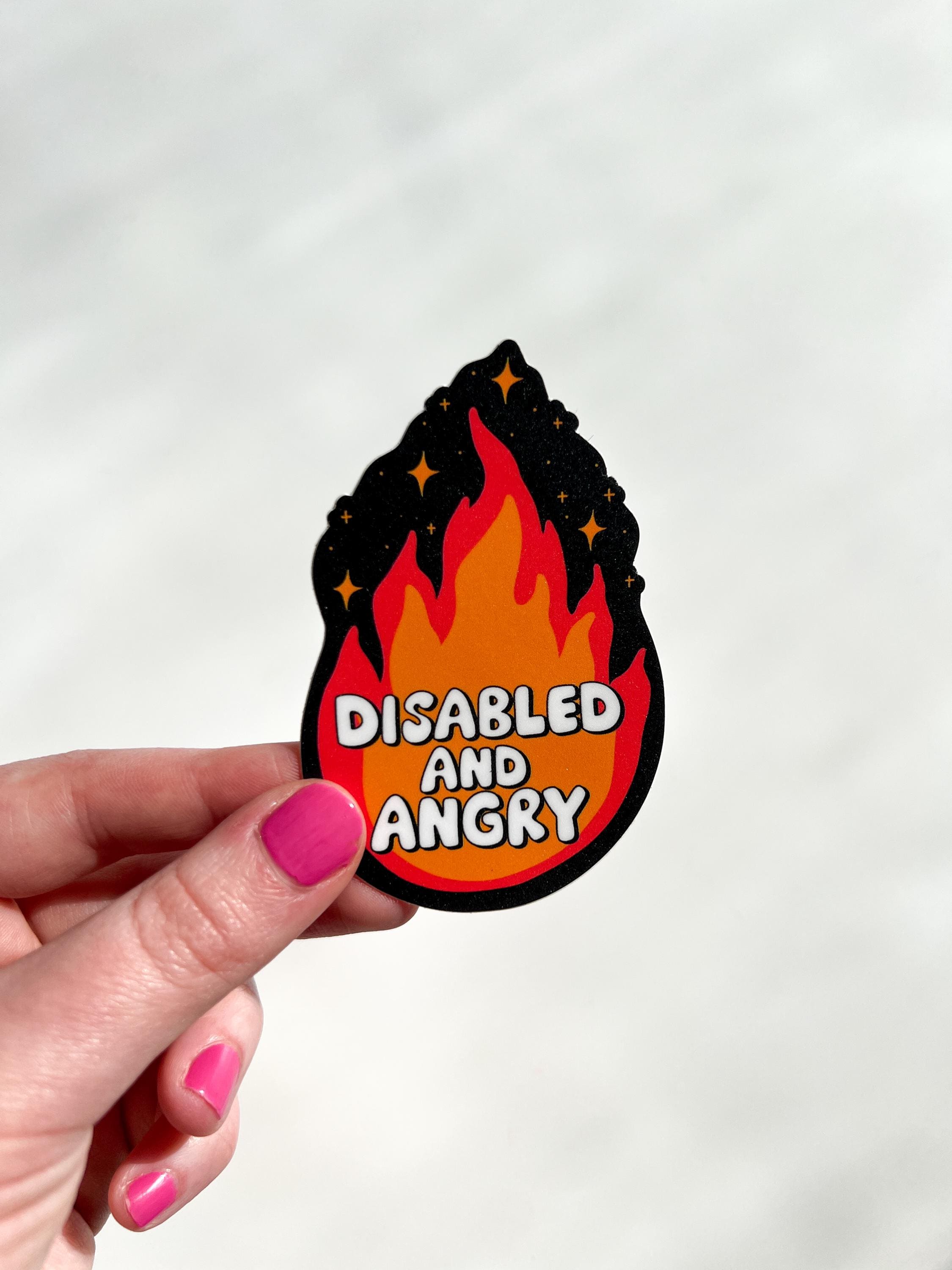 Flame shaped sticker with black background, an orange and red flame with sparkles around the top. The font in the flames center reads, "disabled and angry". The sticker is held in front of white table.
