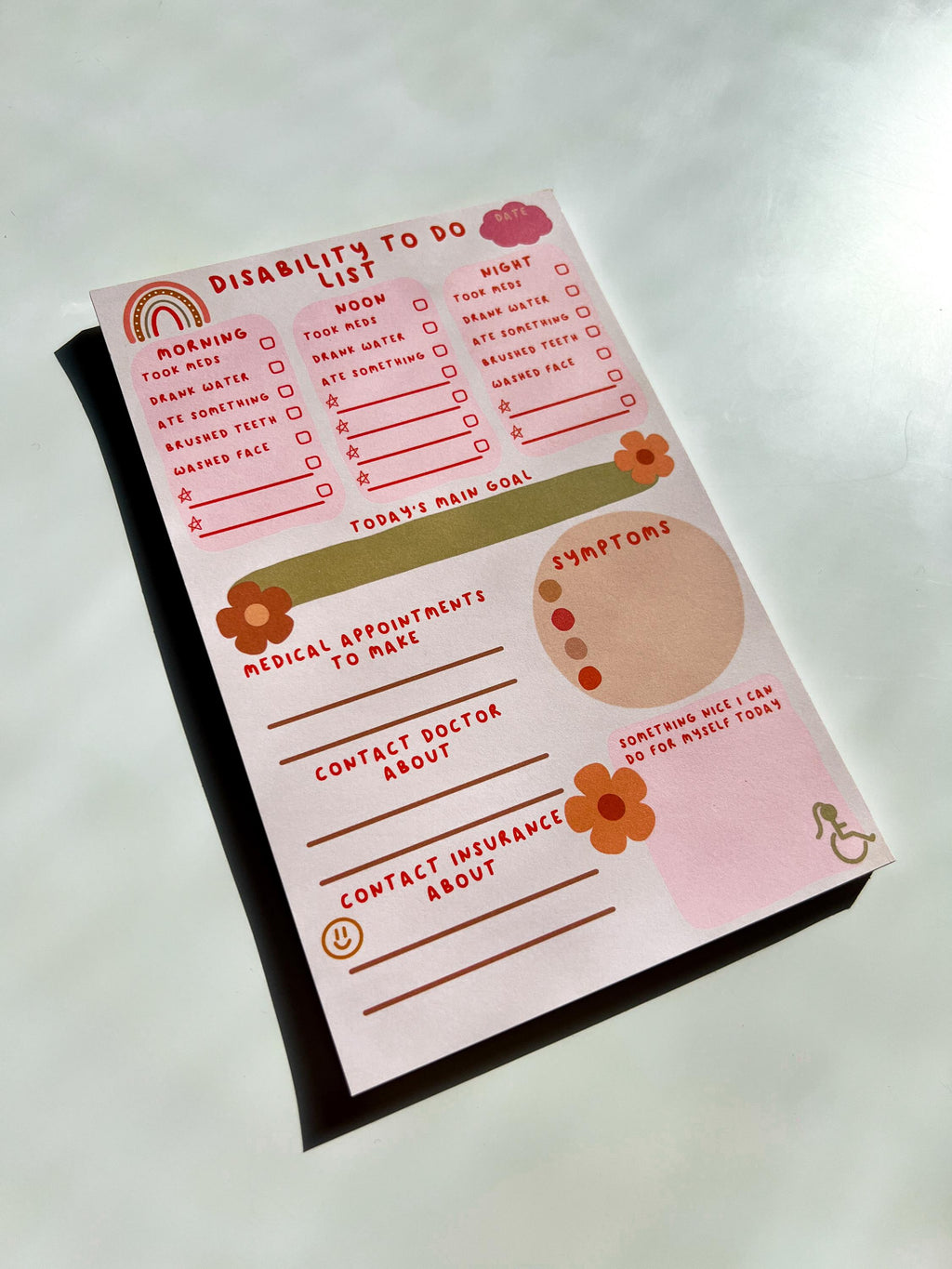 Disability To Do List Notepad with light colored sheets. Sections include: pink cloud to write the date down in, three pink boxes with red font reading "morning, noon, and night" with a checklist underneath them reading "took meds, drank water, ate something, brushed teeth, washed face" and a couple of empty spaces below to fill out yourself. There's a place to write down medical appointments to make, contact doctor about, and contact insurance about. There's a place to track symptoms. On white table