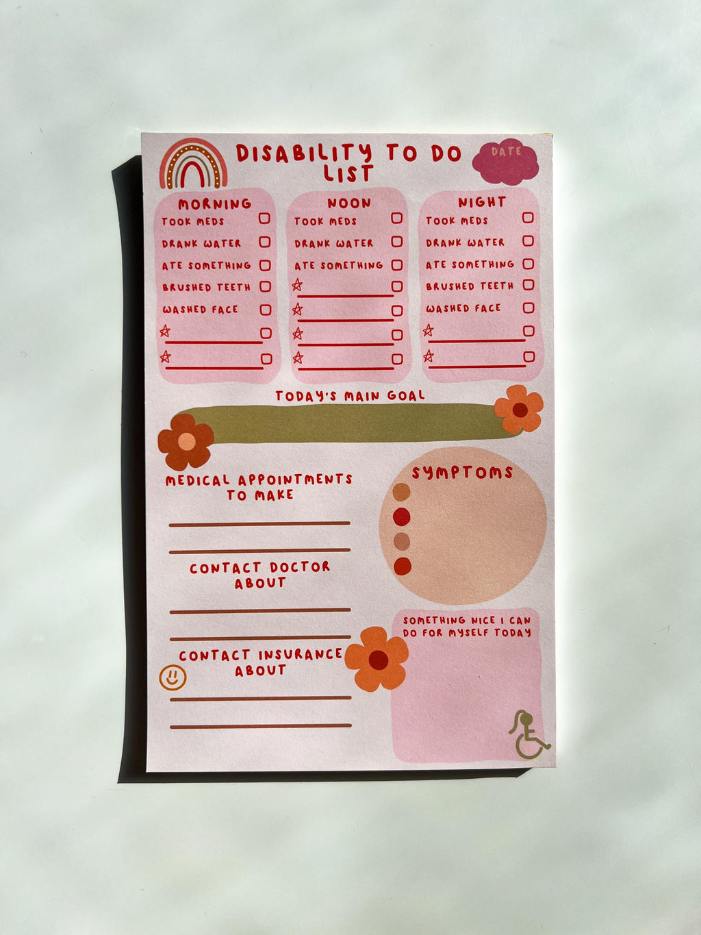 Disability To Do List Notepad with light colored sheets. Sections include: pink cloud to write the date down in, three pink boxes with red font reading "morning, noon, and night" with a checklist underneath them reading "took meds, drank water, ate something, brushed teeth, washed face" and a couple of empty spaces below to fill out yourself. There's a place to write down medical appointments to make, contact doctor about, and contact insurance about. There's a place to track symptoms. On white table
