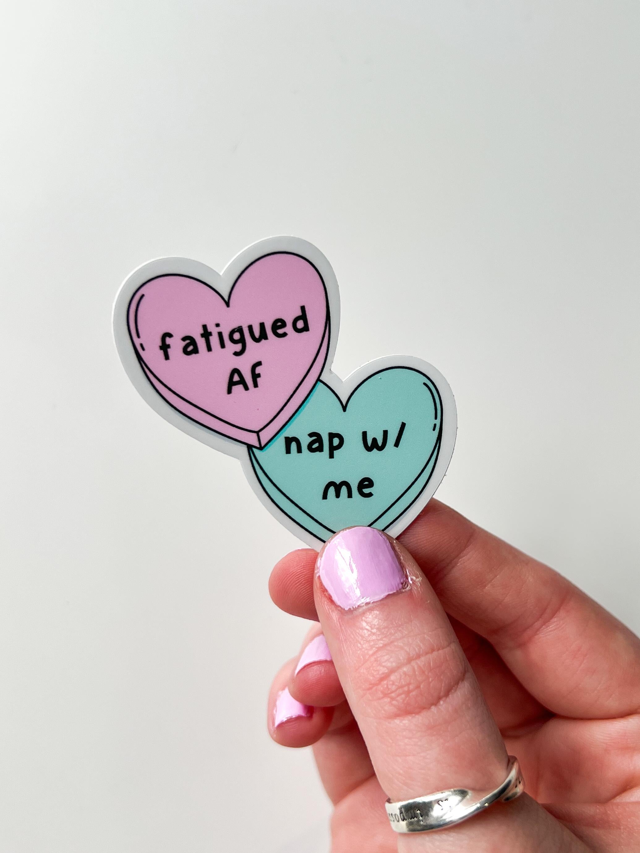Candy heart sticker, one heart is in light pink and the other is in light blue. The font on the pink one reads, "fatigued AF" while the blue heart reads, "nap with me." Sticker is being held over white table. 
