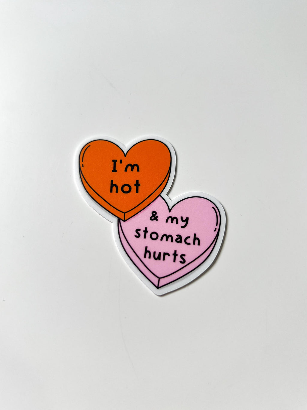 Candy heart sticker with one heart being a bright shade of orange and the other in light pink. The bright orange heart reads, "I'm hot" while the light prink heart reads, "and my stomach hurts". Sticker is laid on white table tilted at an angle. 