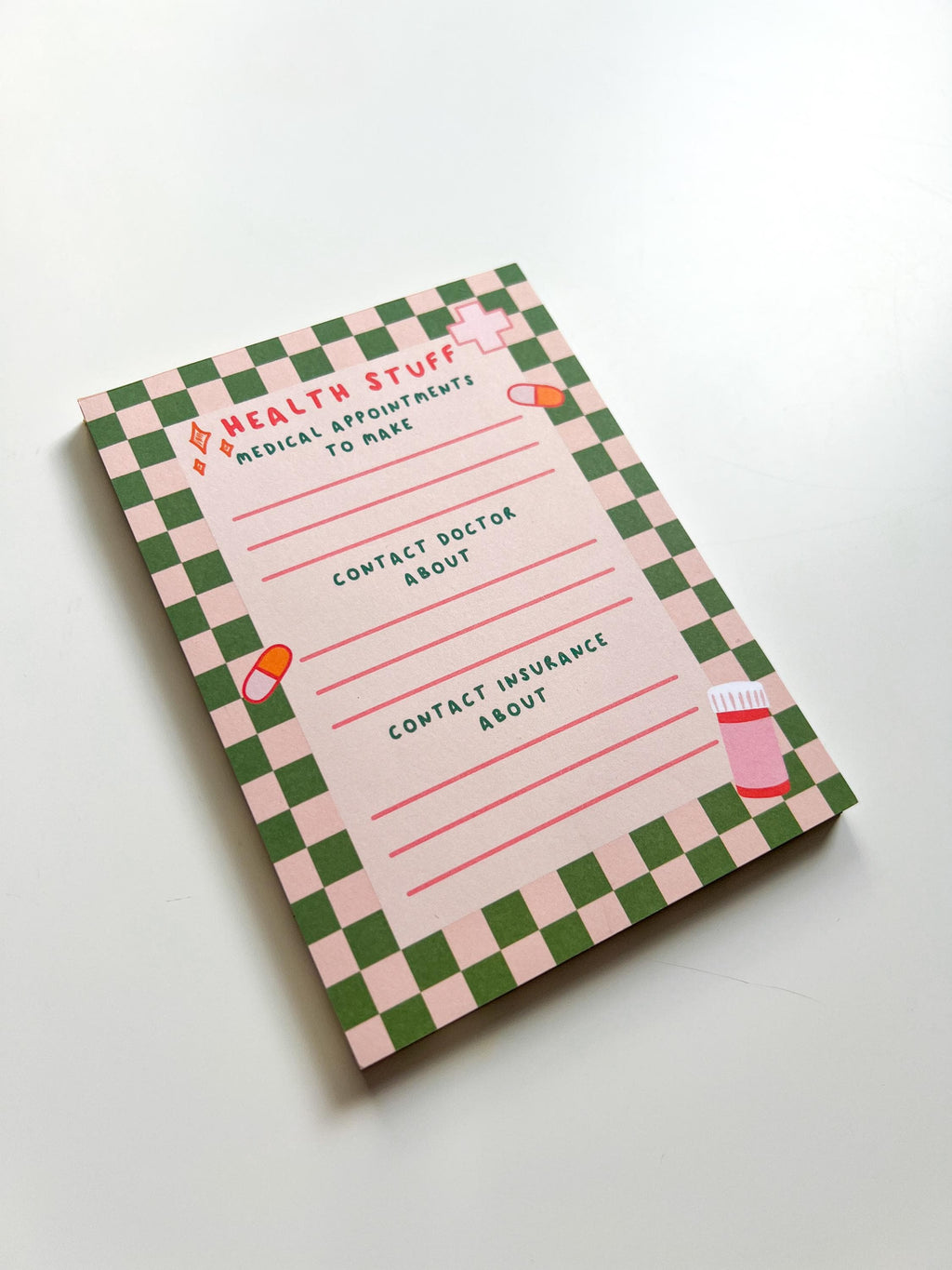 Health To Do List Notepad with beige and bright green checkered print as the border. Top of the page says, "Health Stuff" in a bright pink color. The different sections with pink lines underneath them read, "medical appointments to make, contact doctor about, and contact insurance about". Notepad is pictured laid on white table tilted. 