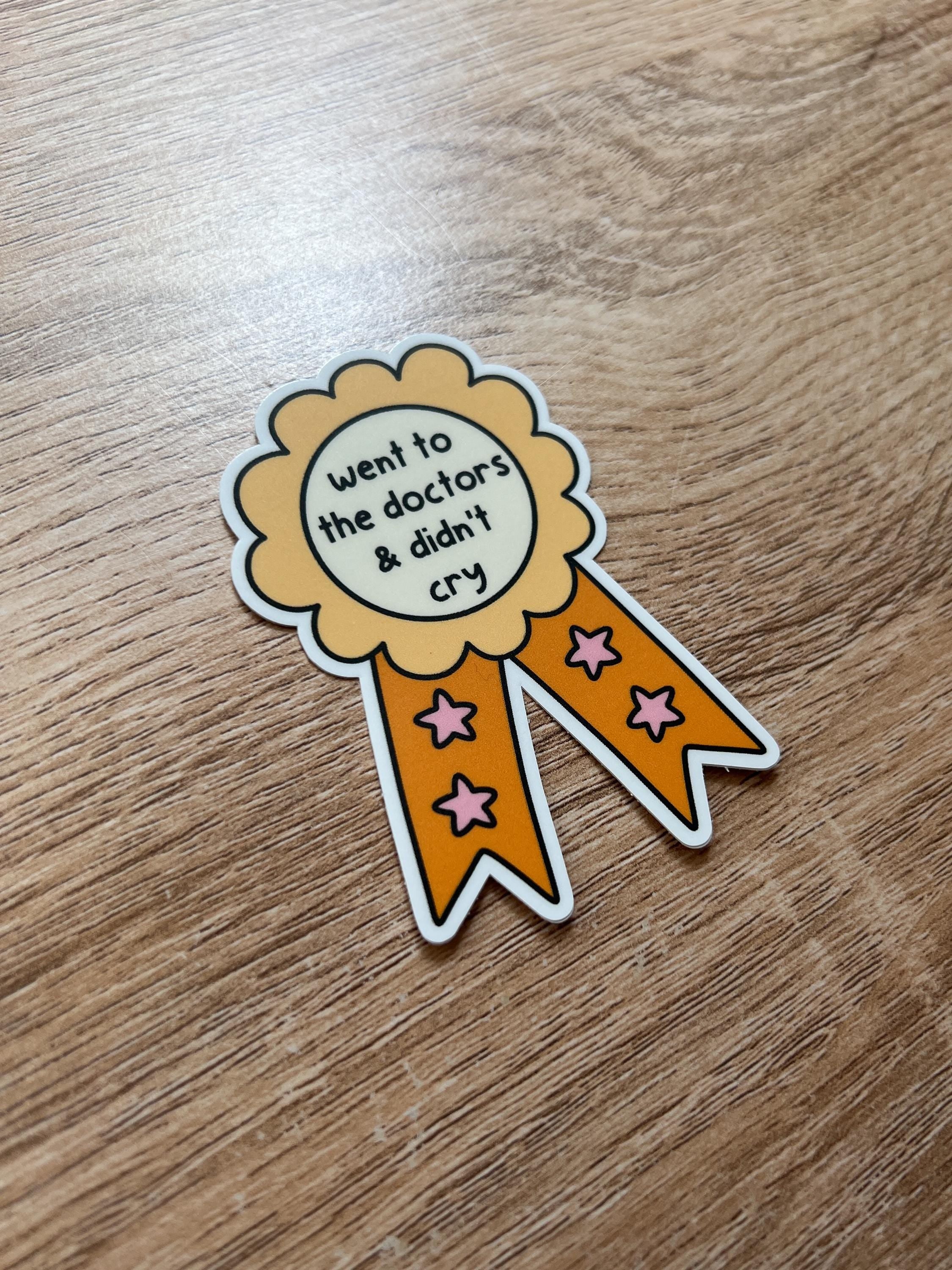 Award ribbon sticker in a light shade of yellow and a bright orange with cream in the center. The black font in the center reads, "went to the doctor's and didn't cry". Sticker is laid on wooden desk tilted to the side. 