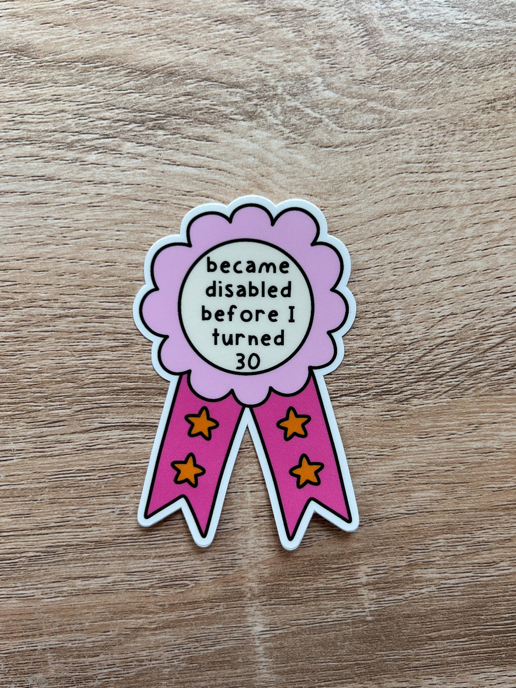 Award ribbon sticker in a light pink and dark pink shades, the middle is cream colored with the black font reading, "became disabled before I turned 30". Sticker is being laid on wooden table. 