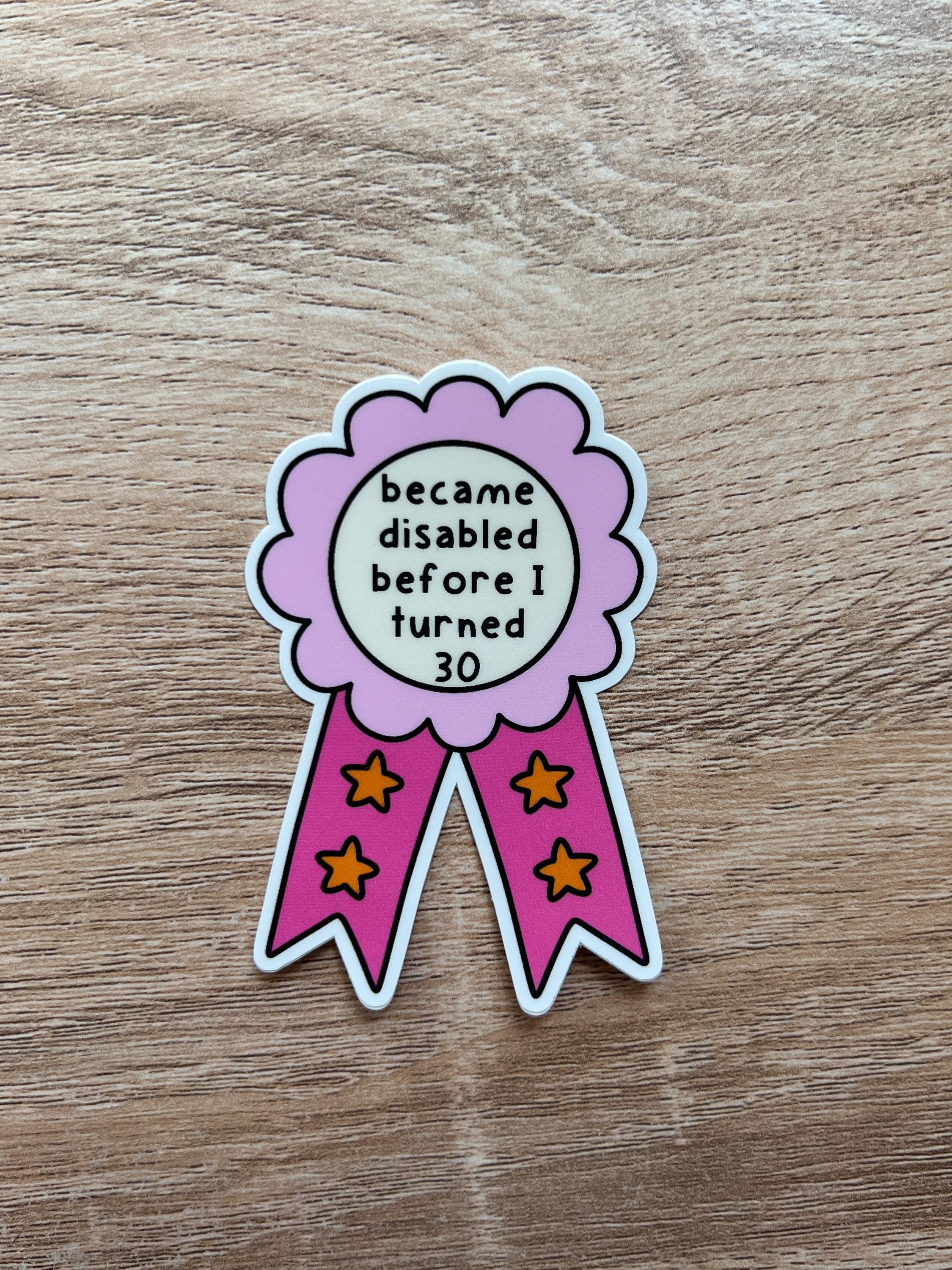 Award ribbon sticker in a light pink and dark pink shades, the middle is cream colored with the black font reading, "became disabled before I turned 30". Sticker is being laid on wooden table. 