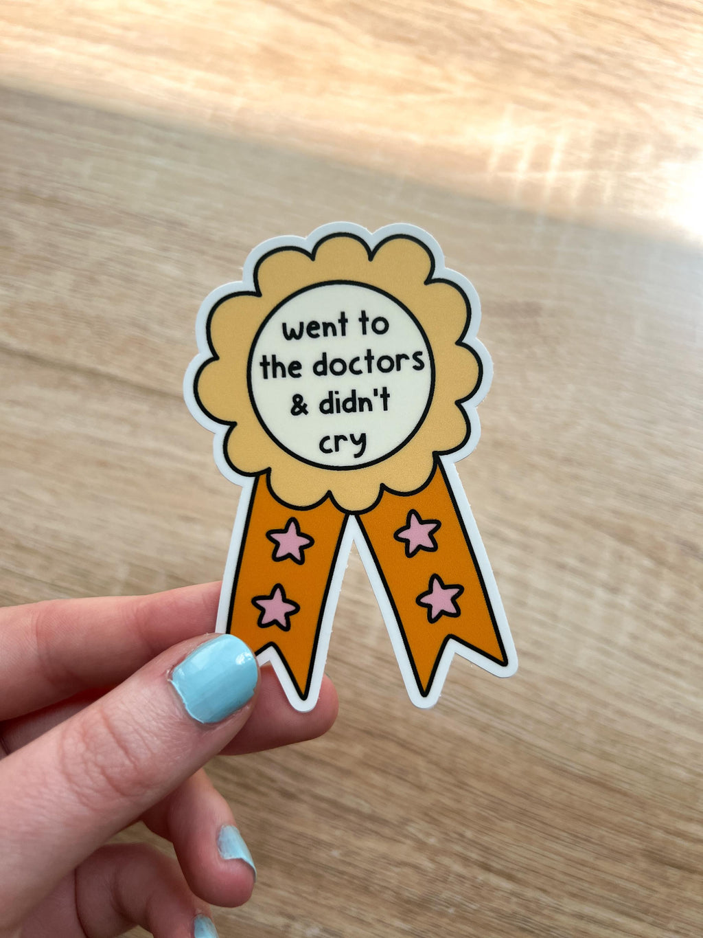 Award ribbon sticker in a light shade of yellow and a bright orange with cream in the center. The black font in the center reads, "went to the doctor's and didn't cry". Sticker is held over wooden desk.