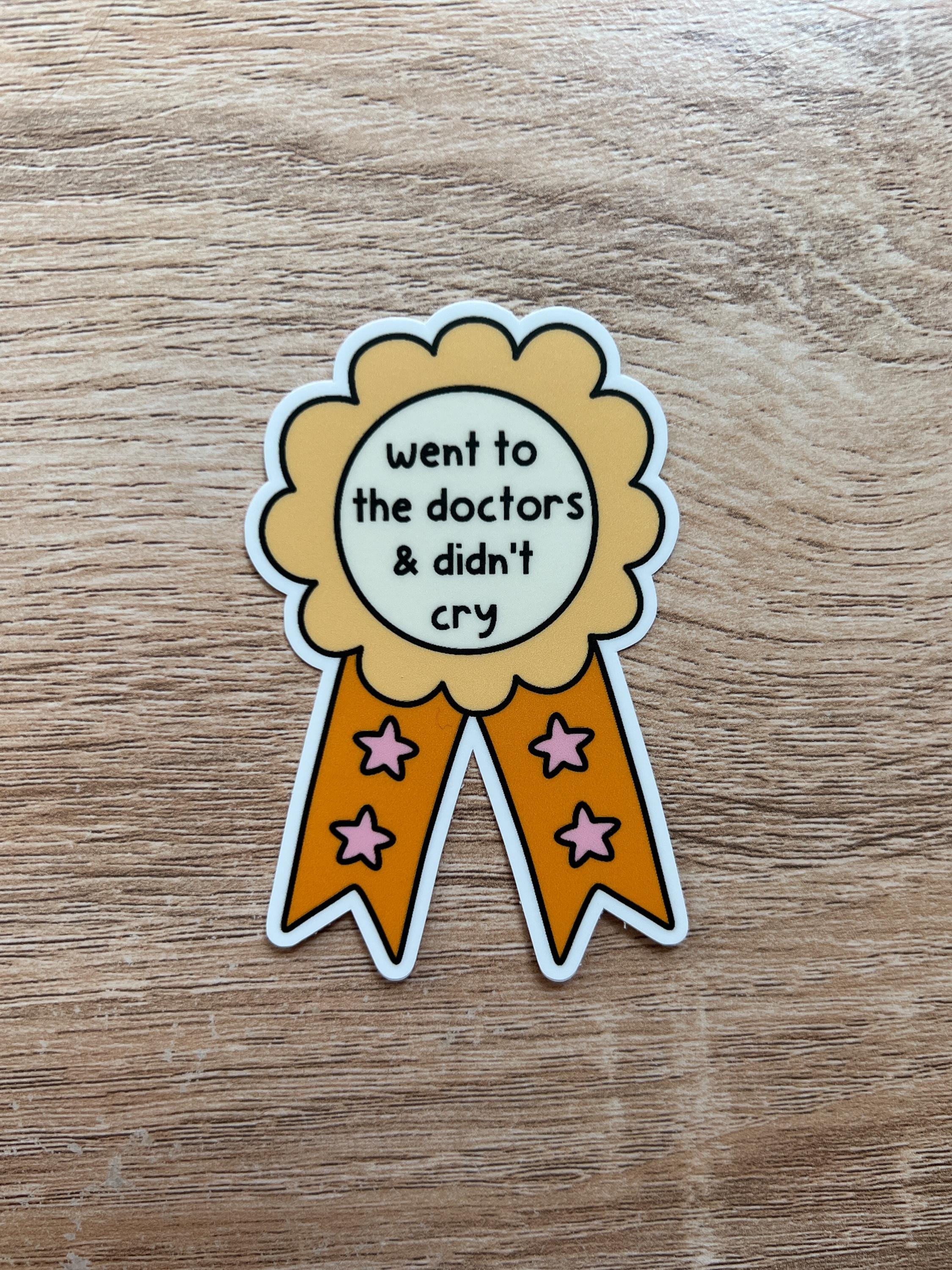 Award ribbon sticker in a light shade of yellow and a bright orange with cream in the center. The black font in the center reads, "went to the doctor's and didn't cry". Sticker is laid on wooden desk. 