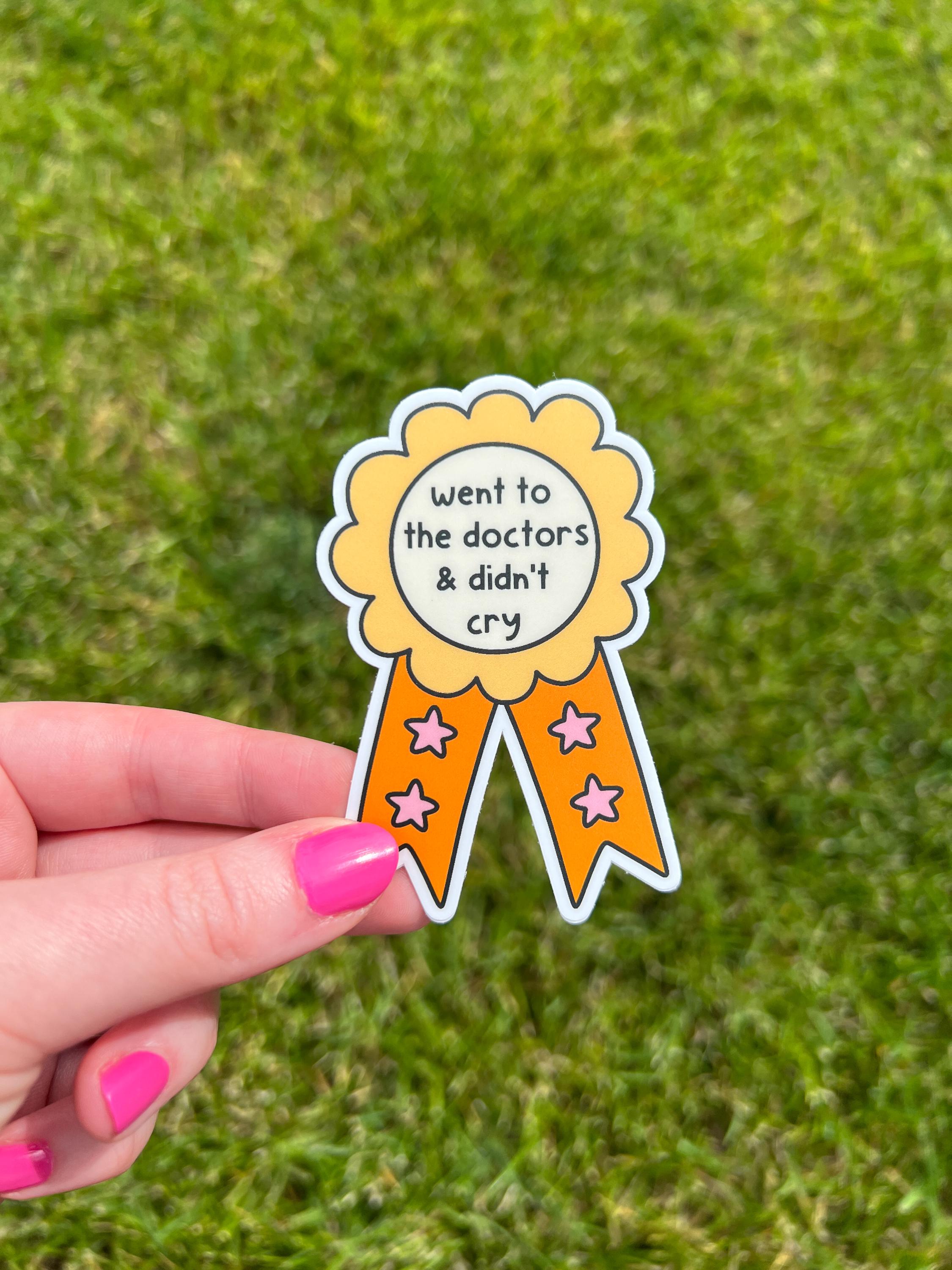 Award ribbon sticker in a light shade of yellow and a bright orange with cream in the center. The black font in the center reads, "went to the doctor's and didn't cry". Sticker is held over grass on a sunny day. 