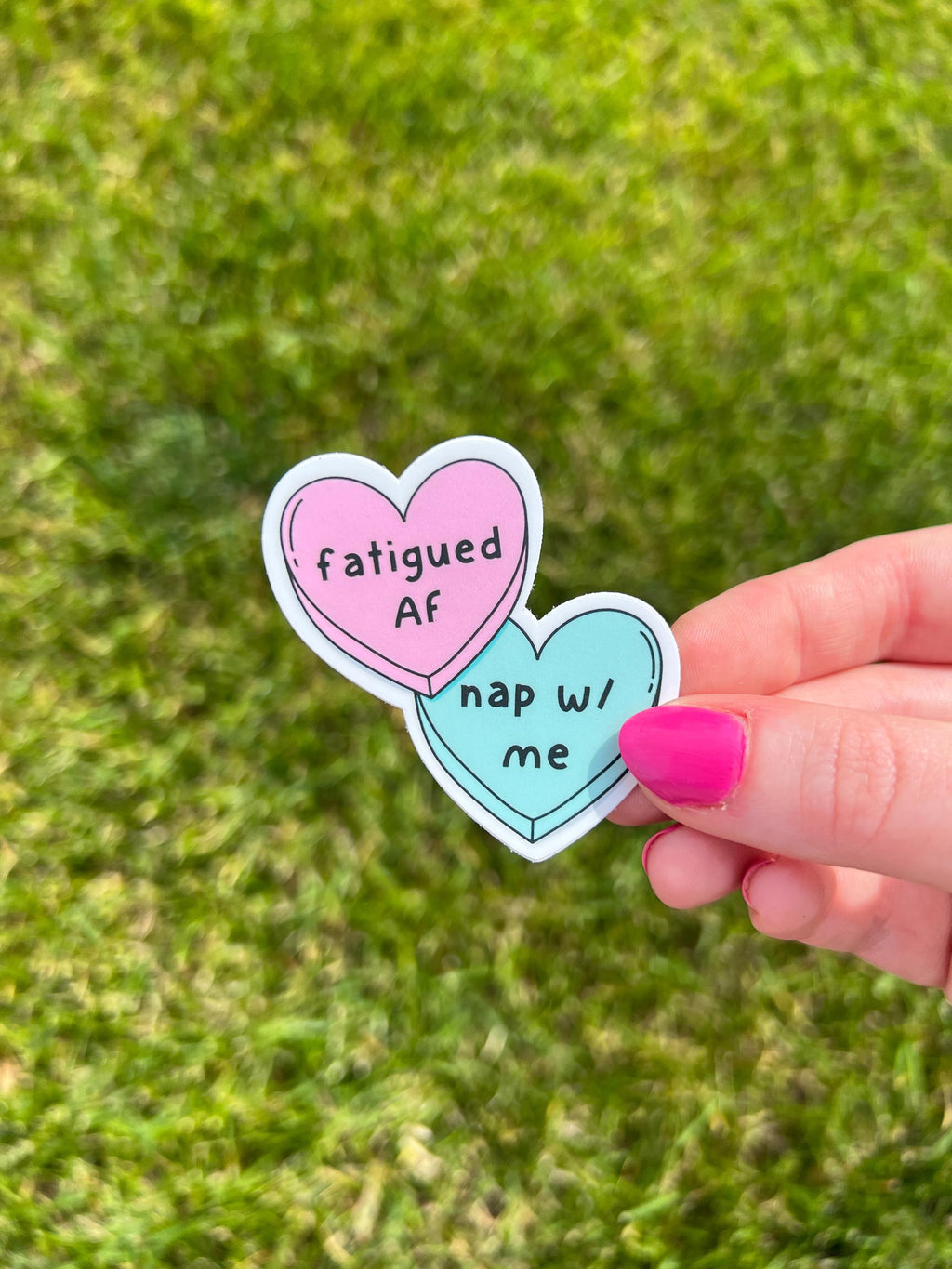 Candy heart sticker, one heart is in light pink and the other is in light blue. The font on the pink one reads, "fatigued AF" while the blue heart reads, "nap with me." Sticker is being held over grass on a sunny day. 