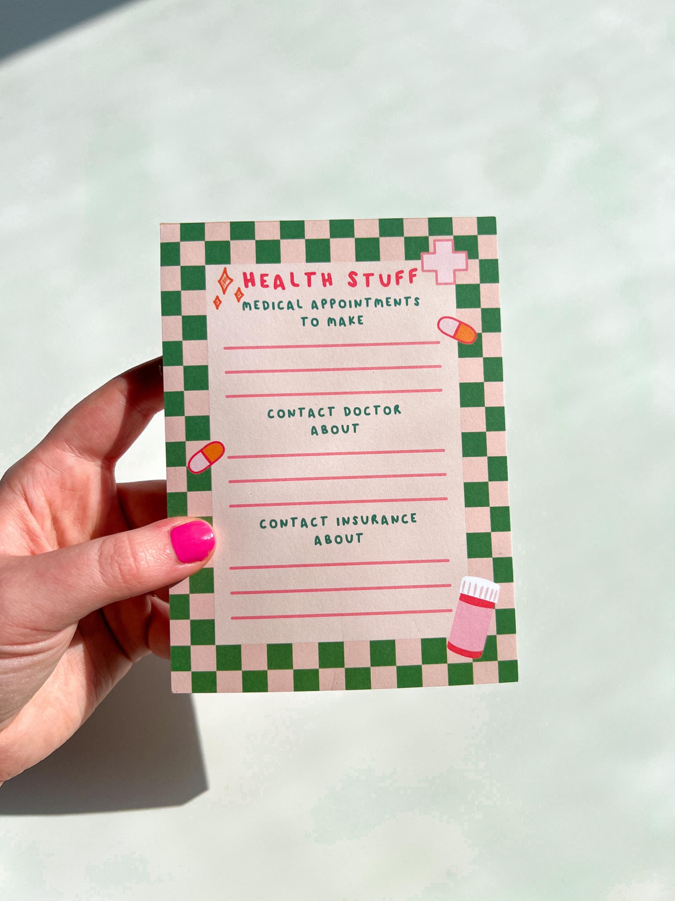 Health To Do List Notepad with beige and bright green checkered print as the border. Top of the page says, "Health Stuff" in a bright pink color. The different sections with pink lines underneath them read, "medical appointments to make, contact doctor about, and contact insurance about". Notepad is pictured being held in front of white table. 