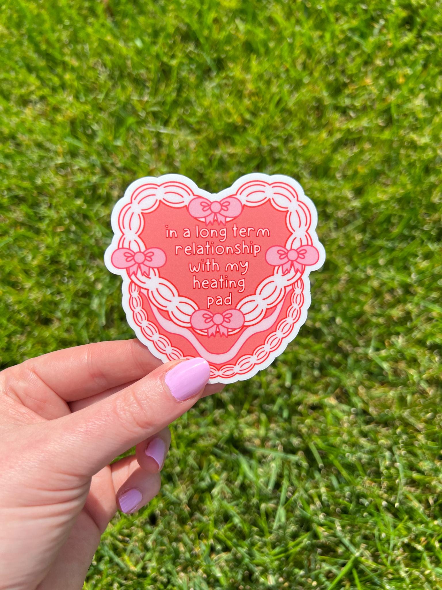 Heart cake shaped sticker. Heart cake is a bright coral color decorated with white frosting and pink bows around the front of the cake. The text outlined in dark red and filled in with white reads, "in a long term relationship with my heating pad". Sticker is held over grass. 